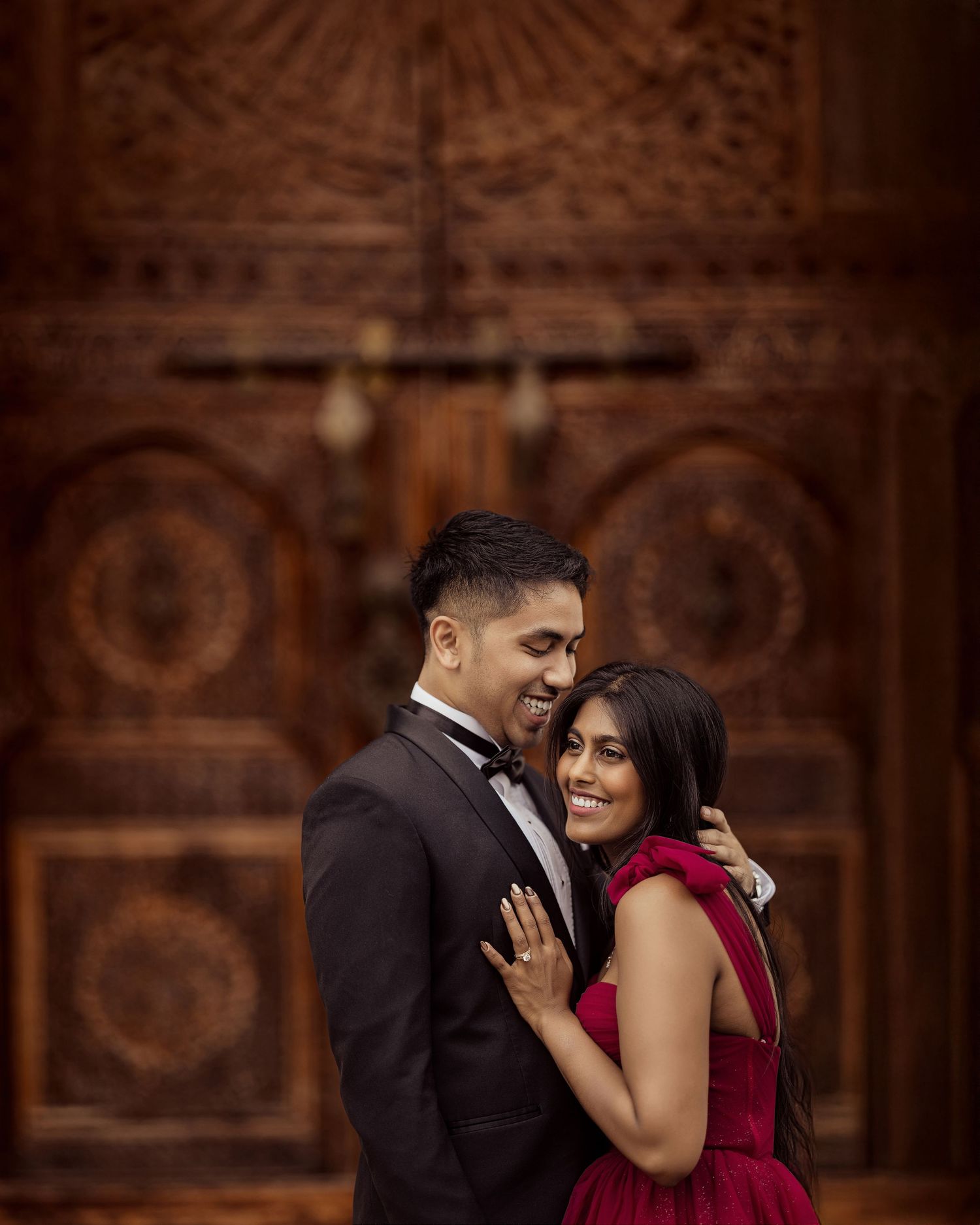 A couple in formal attire embracing in a romantic moment against an ornate dark wooden backdrop.
