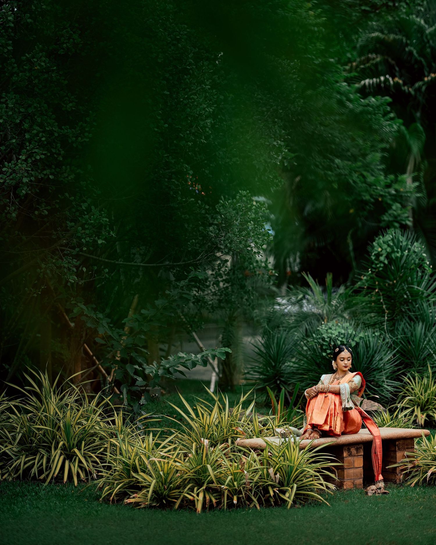 A romantic couple in rust-colored outfits sits on a stone bench surrounded by lush tropical garden foliage.