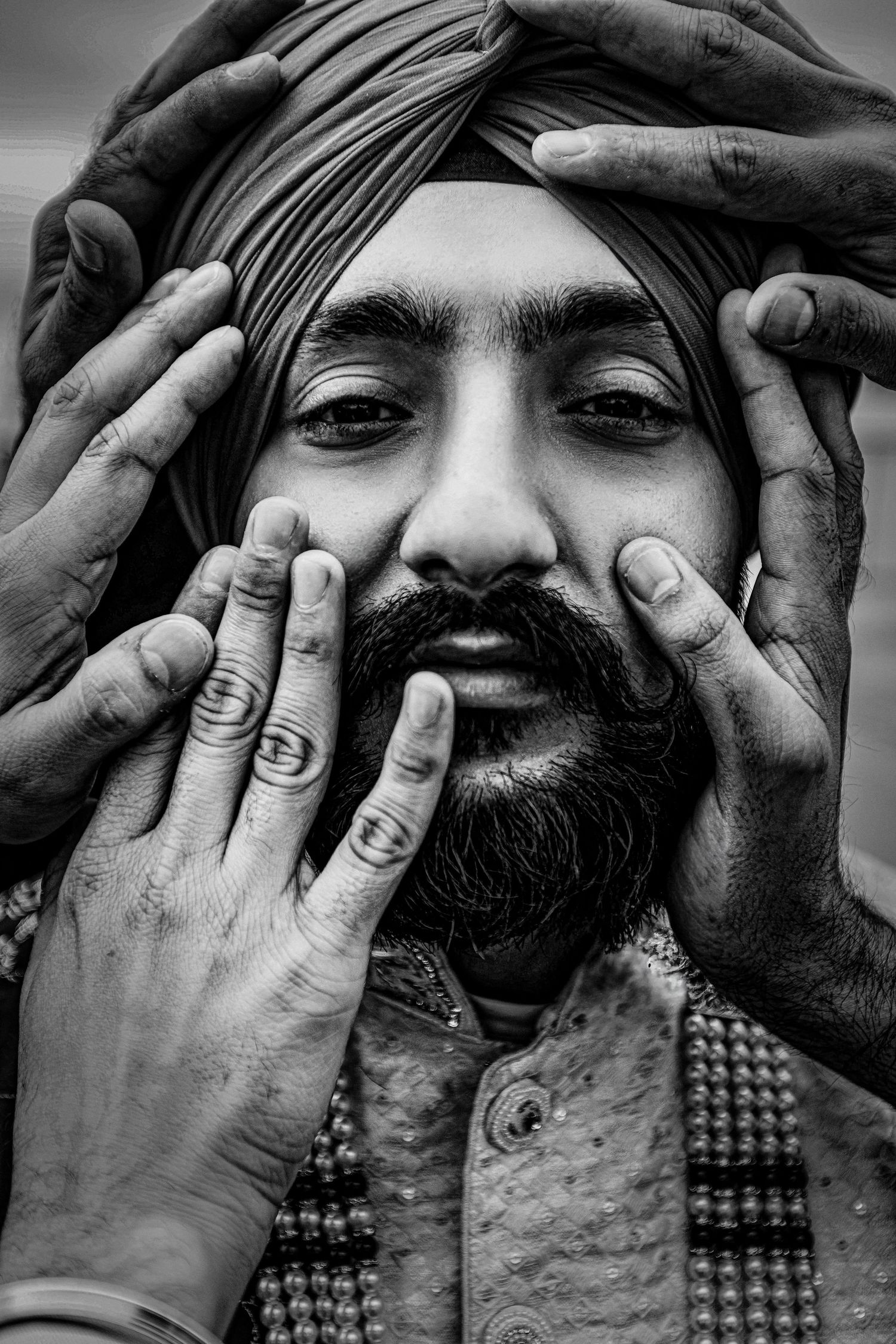 Black and white portrait of a Sikh man in turban with multiple hands gently framing his bearded face.