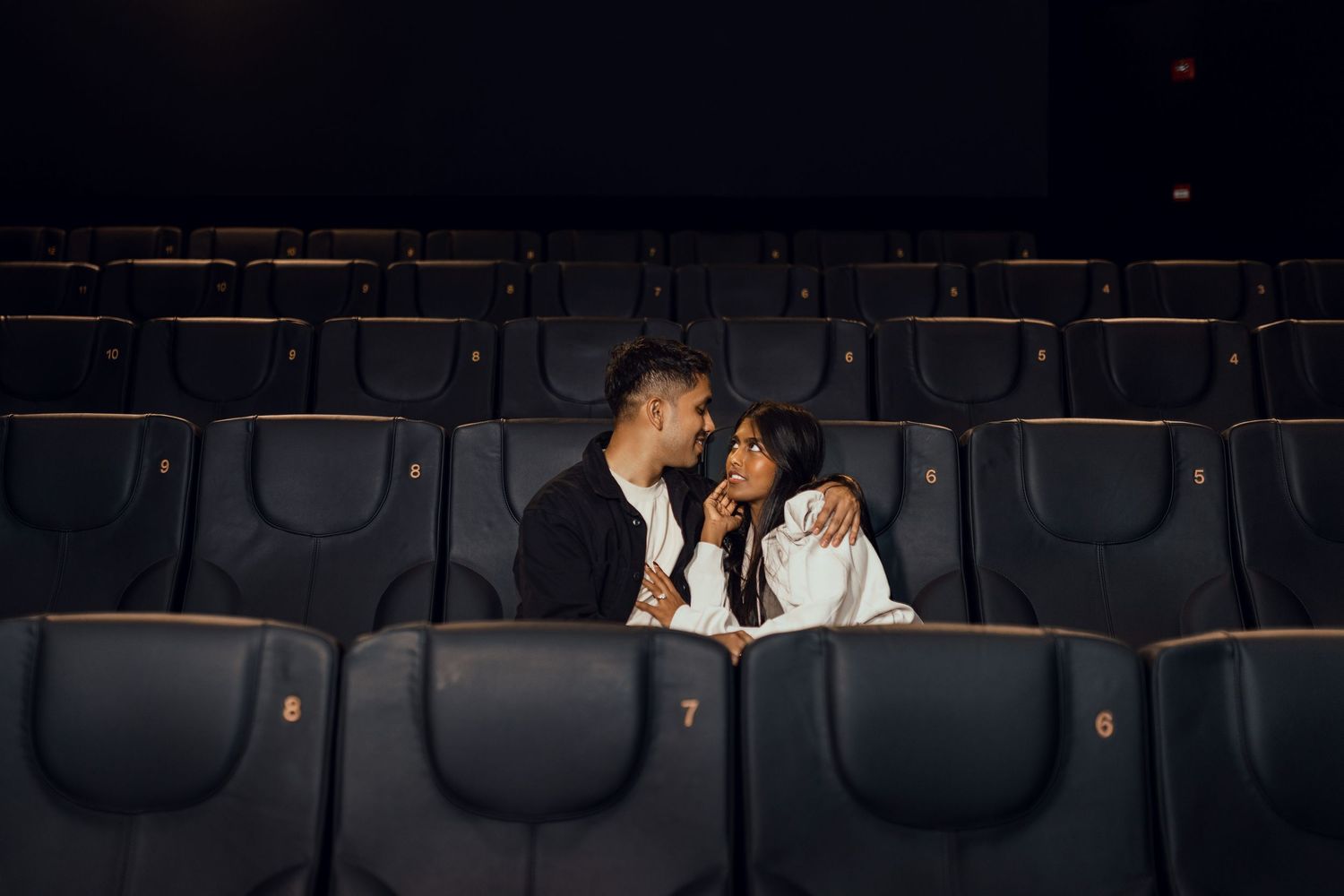 A romantic couple shares an intimate moment alone in an empty dark movie theater, sitting close together in blue seats.