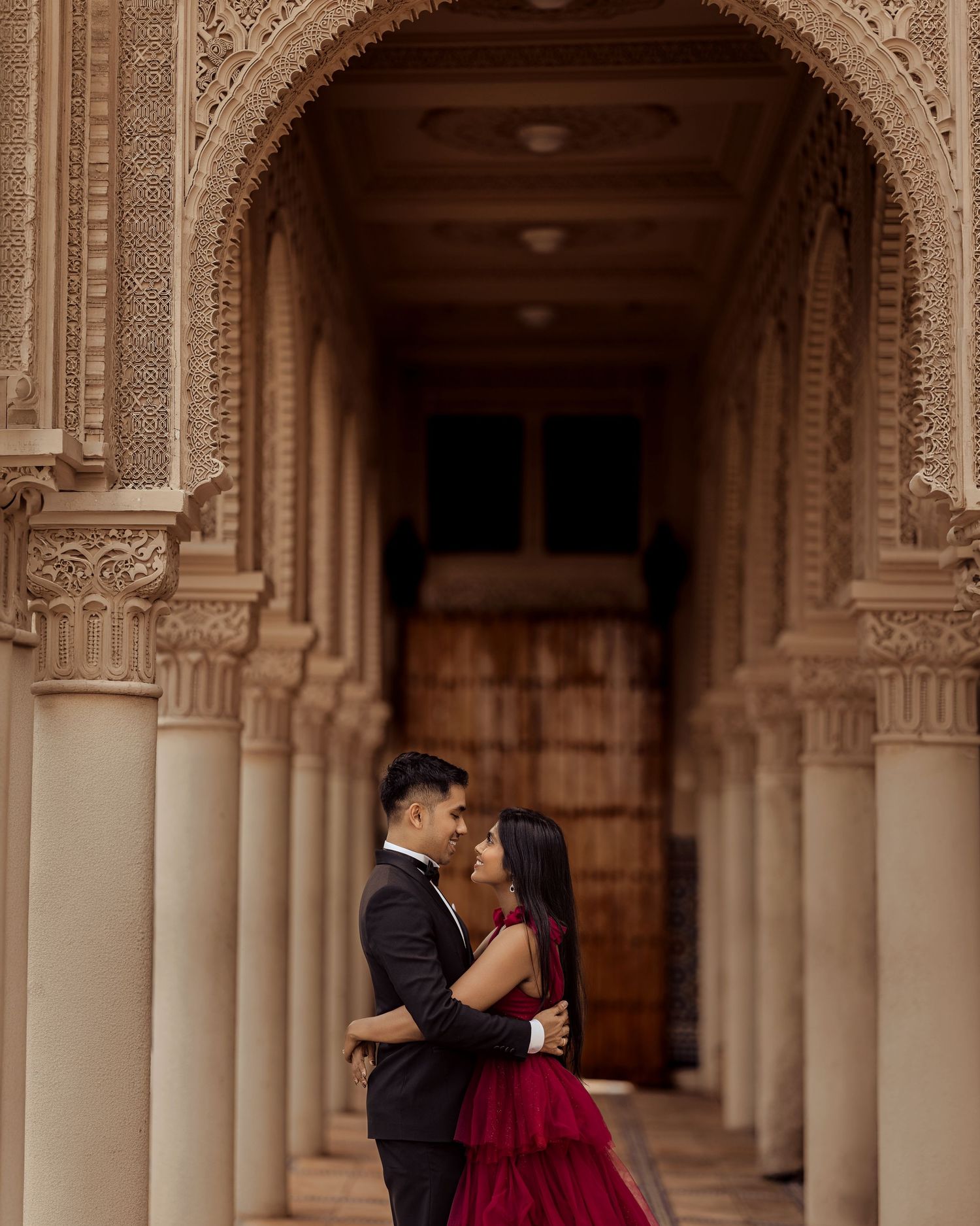 Couple embracing romantically under ornate Moorish arched colonnade, woman in red gown and man in black suit.