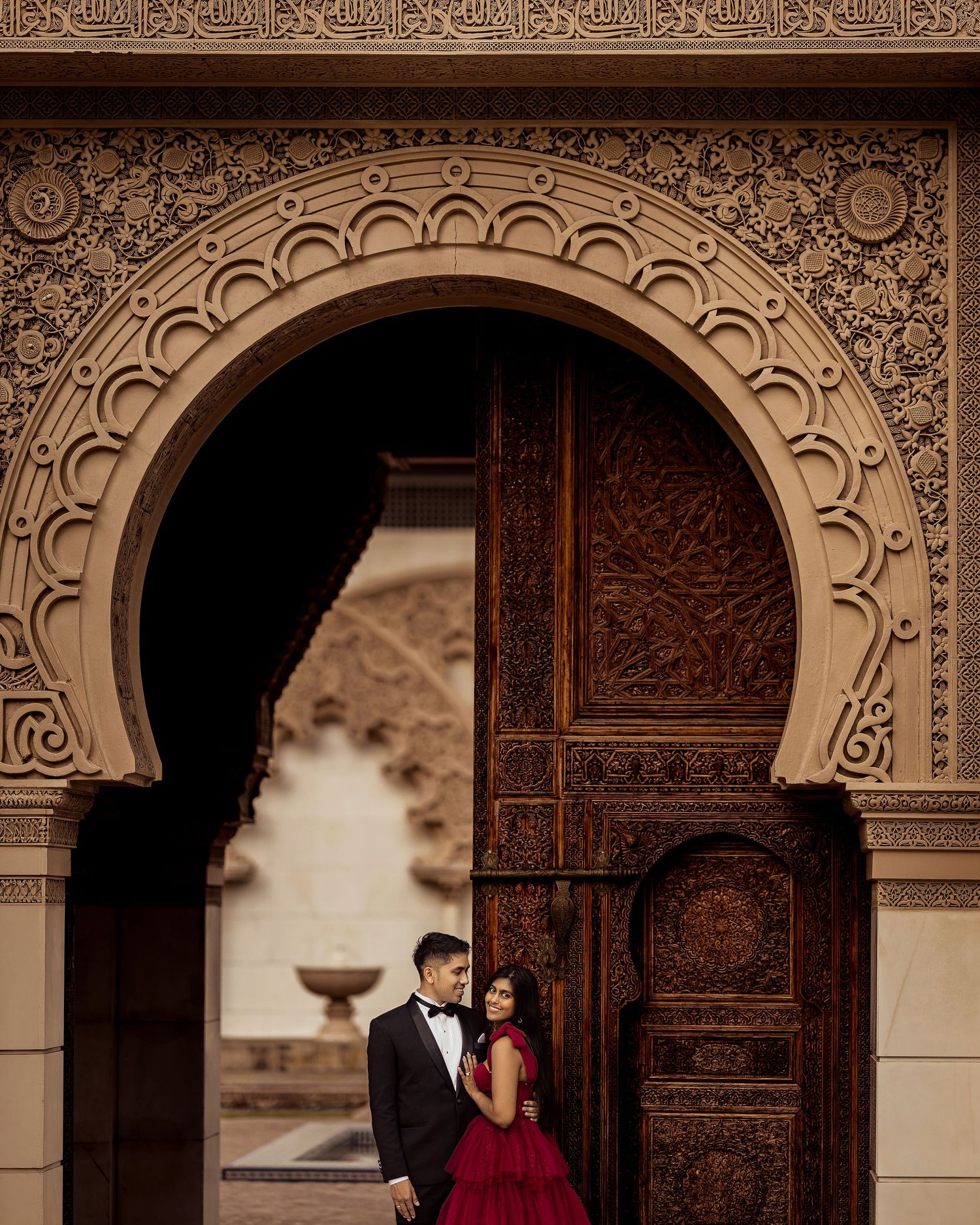 Couple in elegant attire poses under ornate Moroccan arch with intricate carvings and large wooden door.