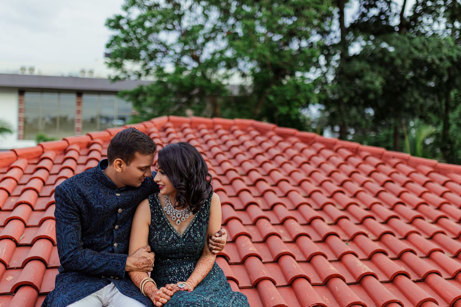 A couple sits together on a red tile roof, sharing an intimate moment on a cloudy day.