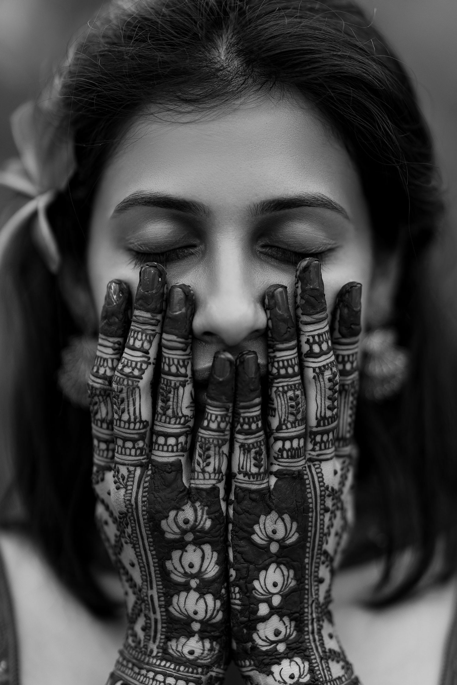 Black and white artistic portrait showing intricate henna designs of lotus flowers on hands covering face.
