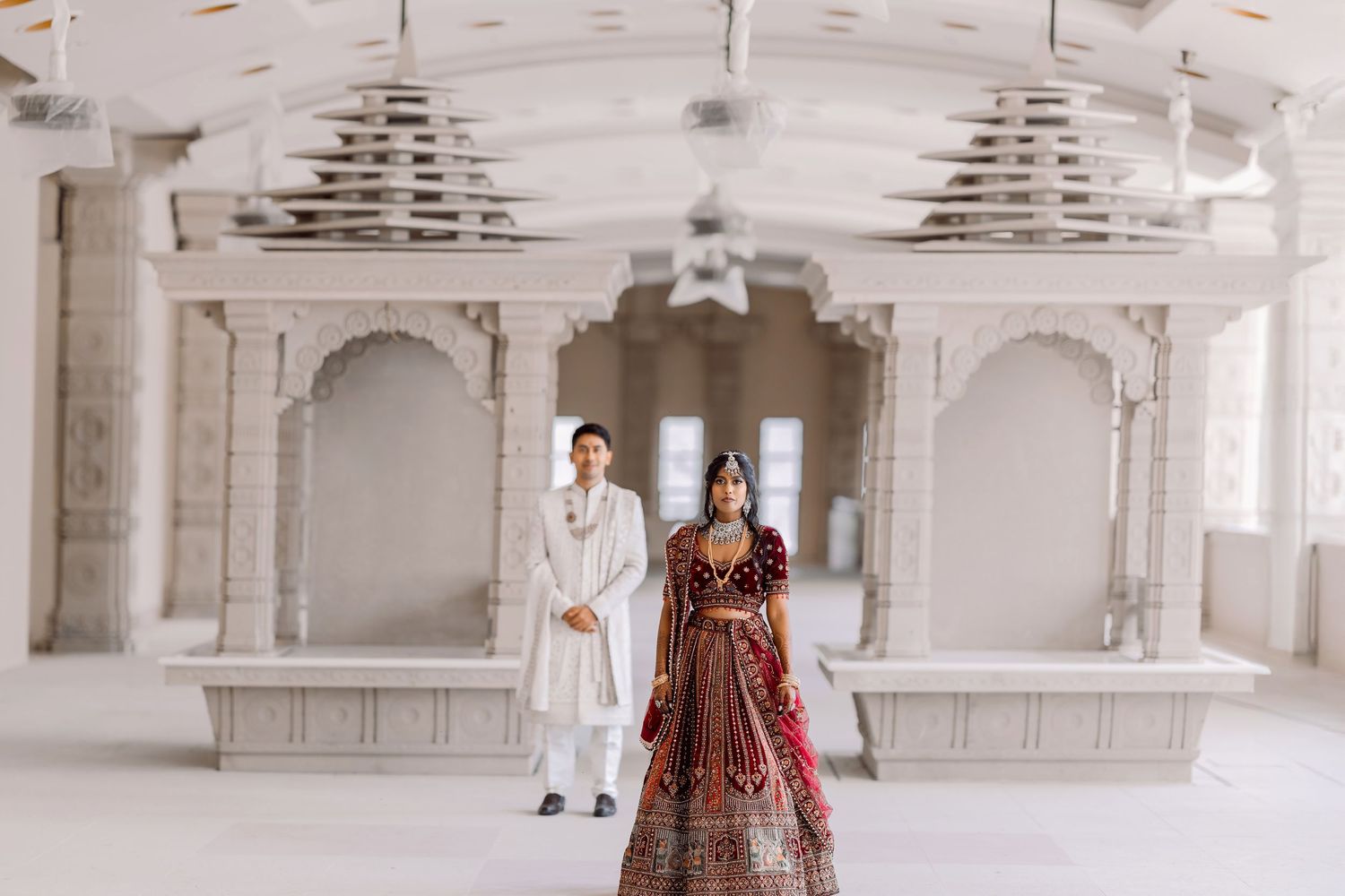 Couple in traditional Indian wedding attire standing inside a white marble temple venue.