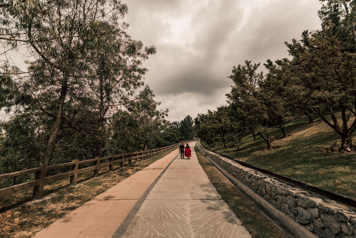 Two people walking on a scenic pathway surrounded by lush green trees under a cloudy sky.