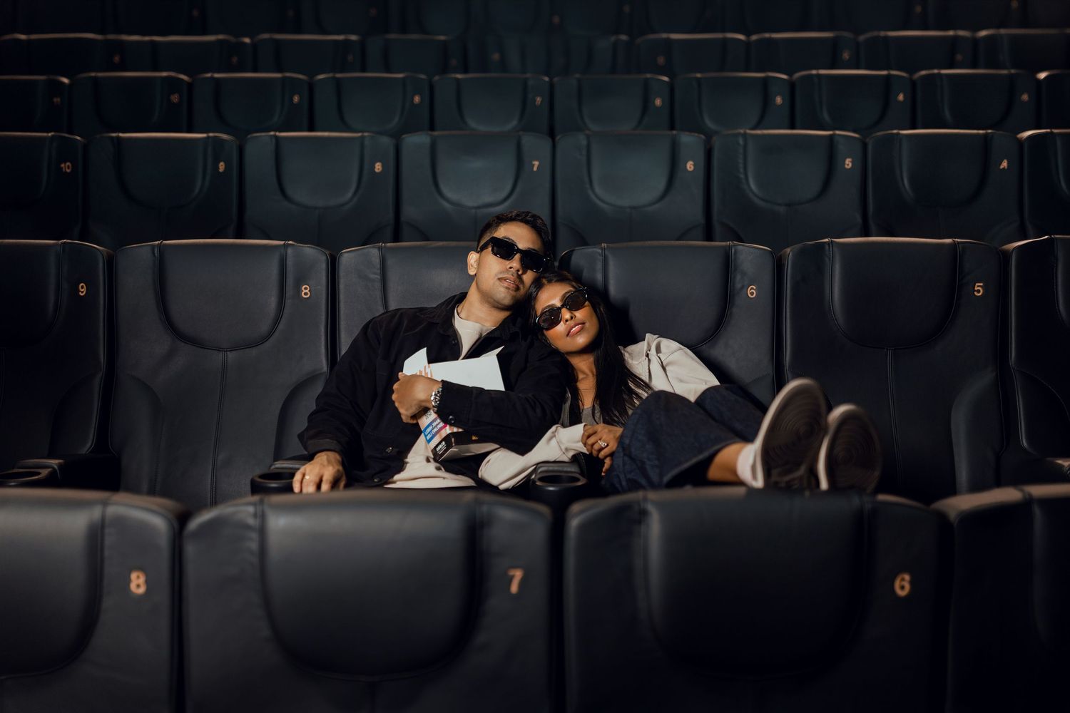 Couple wearing sunglasses cuddling in empty movie theater seats, enjoying a romantic cinema date together.