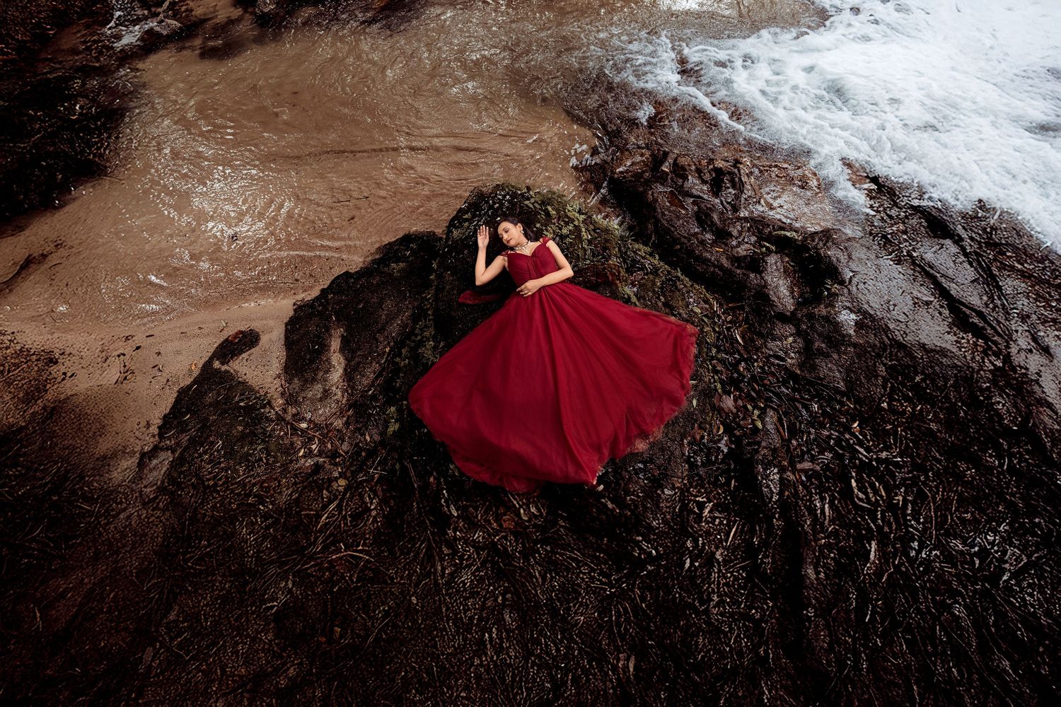 A person in a flowing red dress poses dramatically on dark volcanic rocks near the ocean shore.