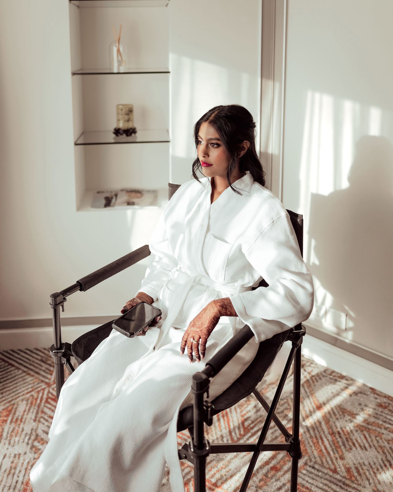 A woman in a white coat dress sits gracefully in a dark chair, bathed in natural light against a minimalist white interior.