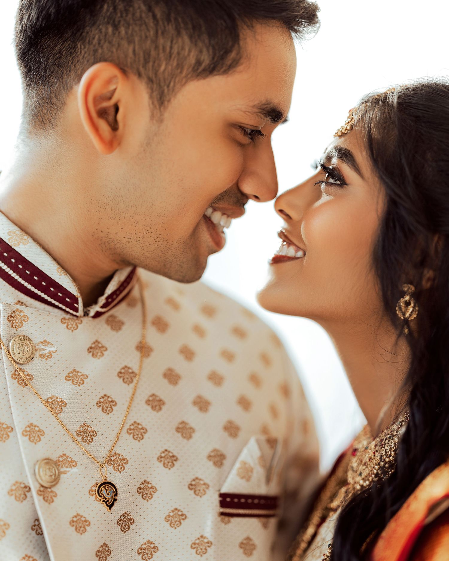 A couple in traditional Indian wedding attire smiling intimately, the groom in an embroidered sherwani and bride in gold jewelry.