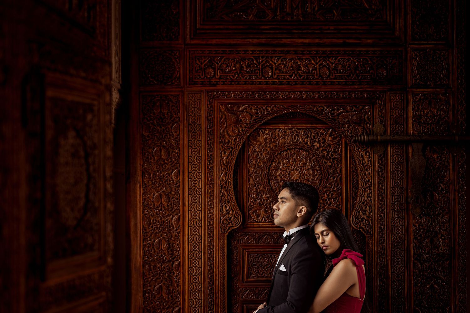 Couple in formal attire embracing against an ornate carved wooden door in warm tones.