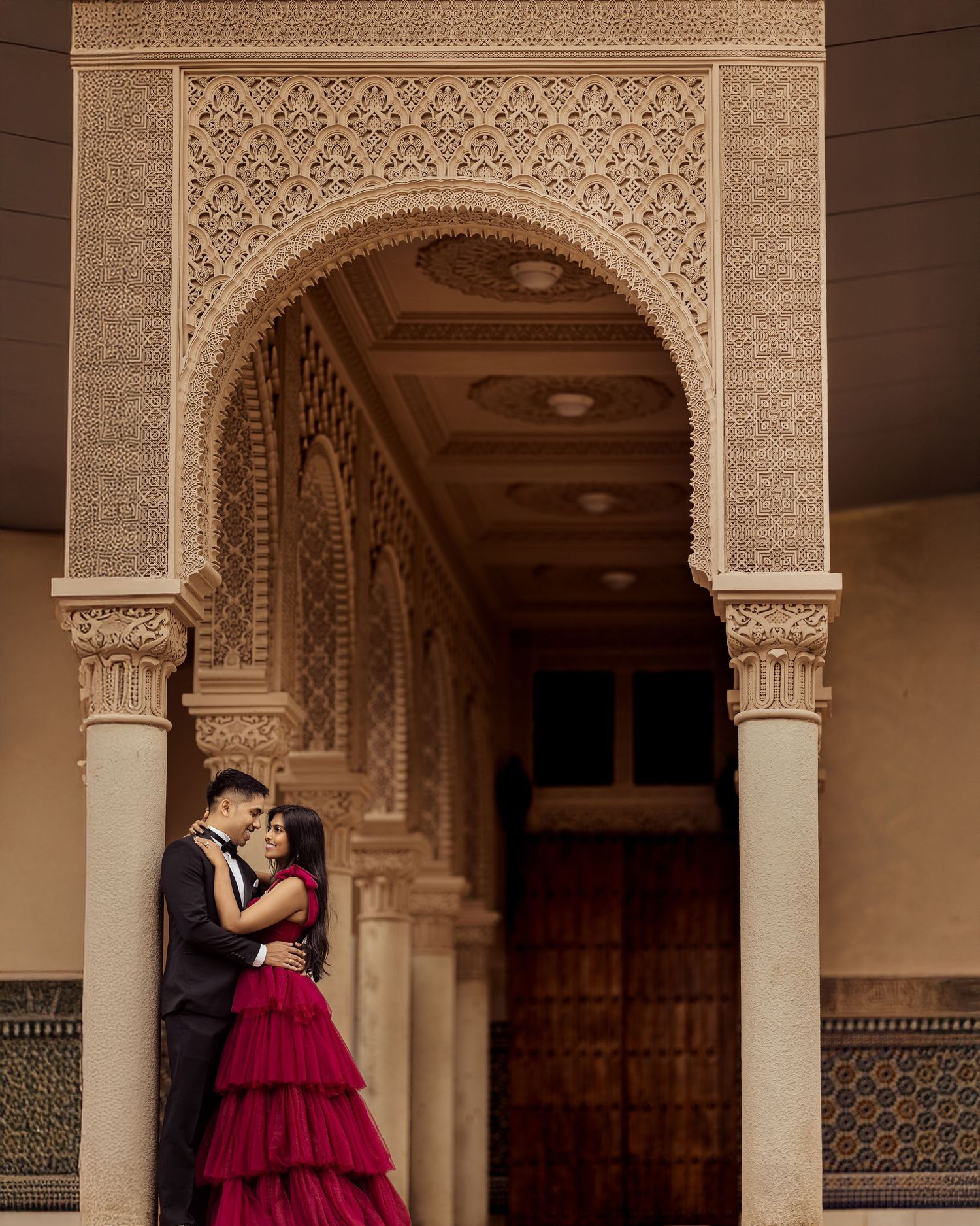 Romantic couple in formal attire embraces under ornate Moorish architecture with intricate arches and tile work.