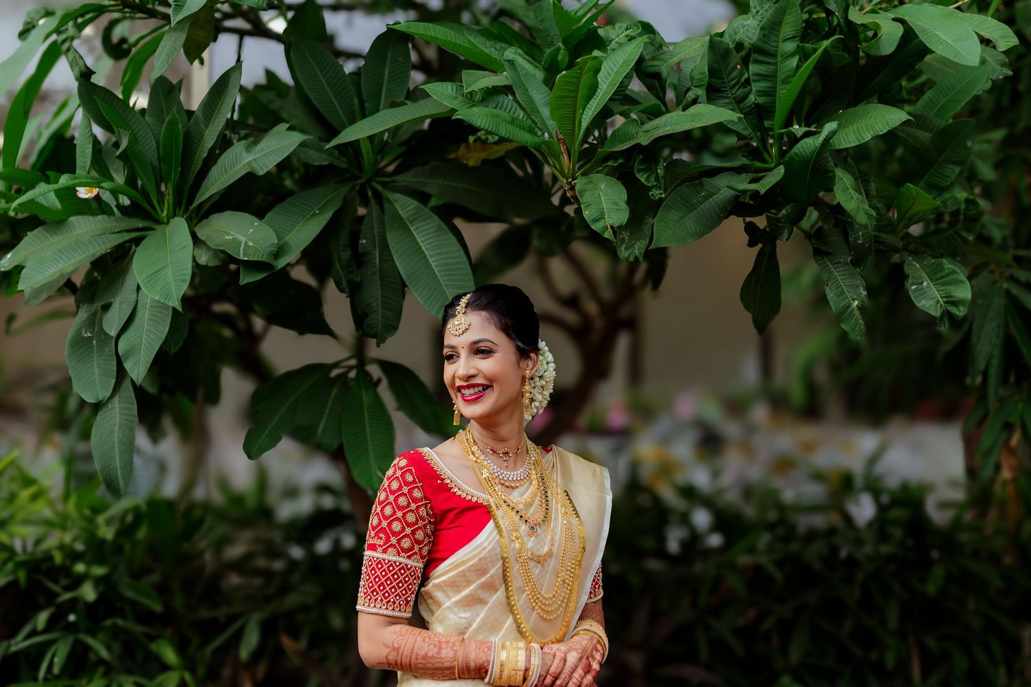 A woman in traditional South Indian bridal attire with gold silk saree and red blouse smiles while seated among green foliage.