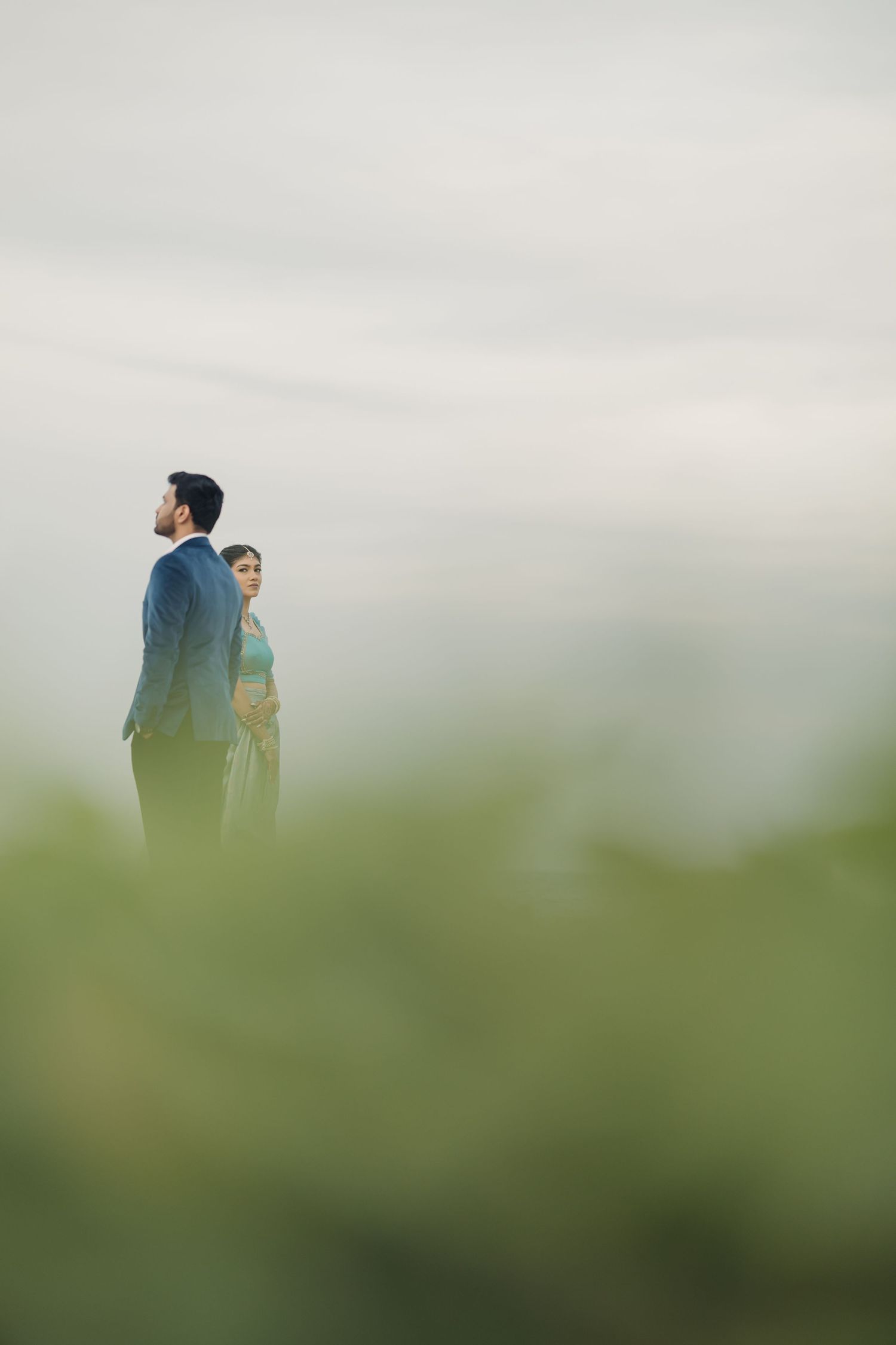 A couple stands back-to-back in a dreamy outdoor setting with soft green bokeh and misty atmosphere.