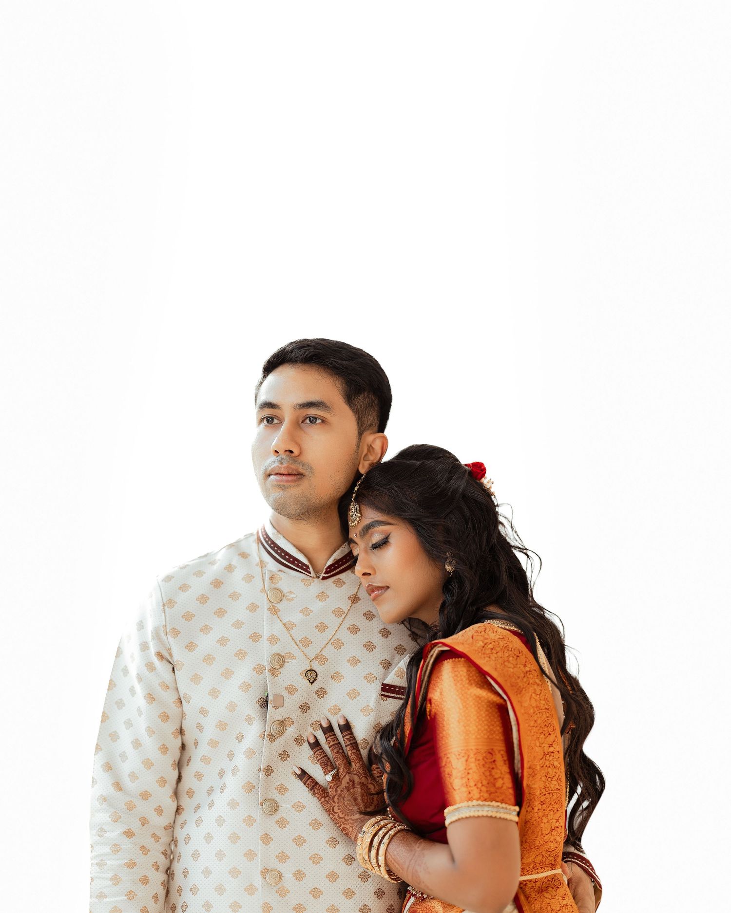Indian couple in traditional wedding attire, man in white sherwani, woman in orange saree with mehndi hands.