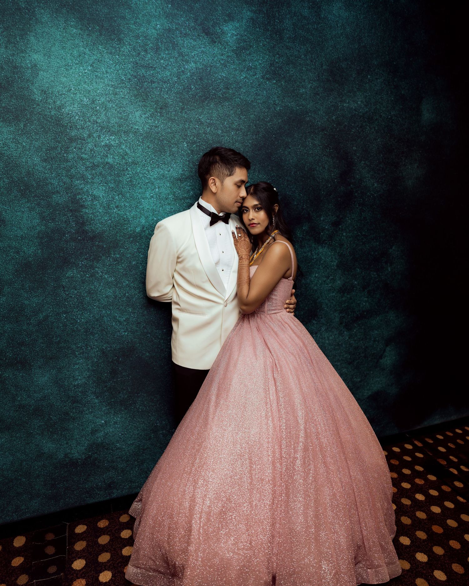 Couple posing elegantly, man in white tuxedo embracing woman in pink ball gown, against teal backdrop.