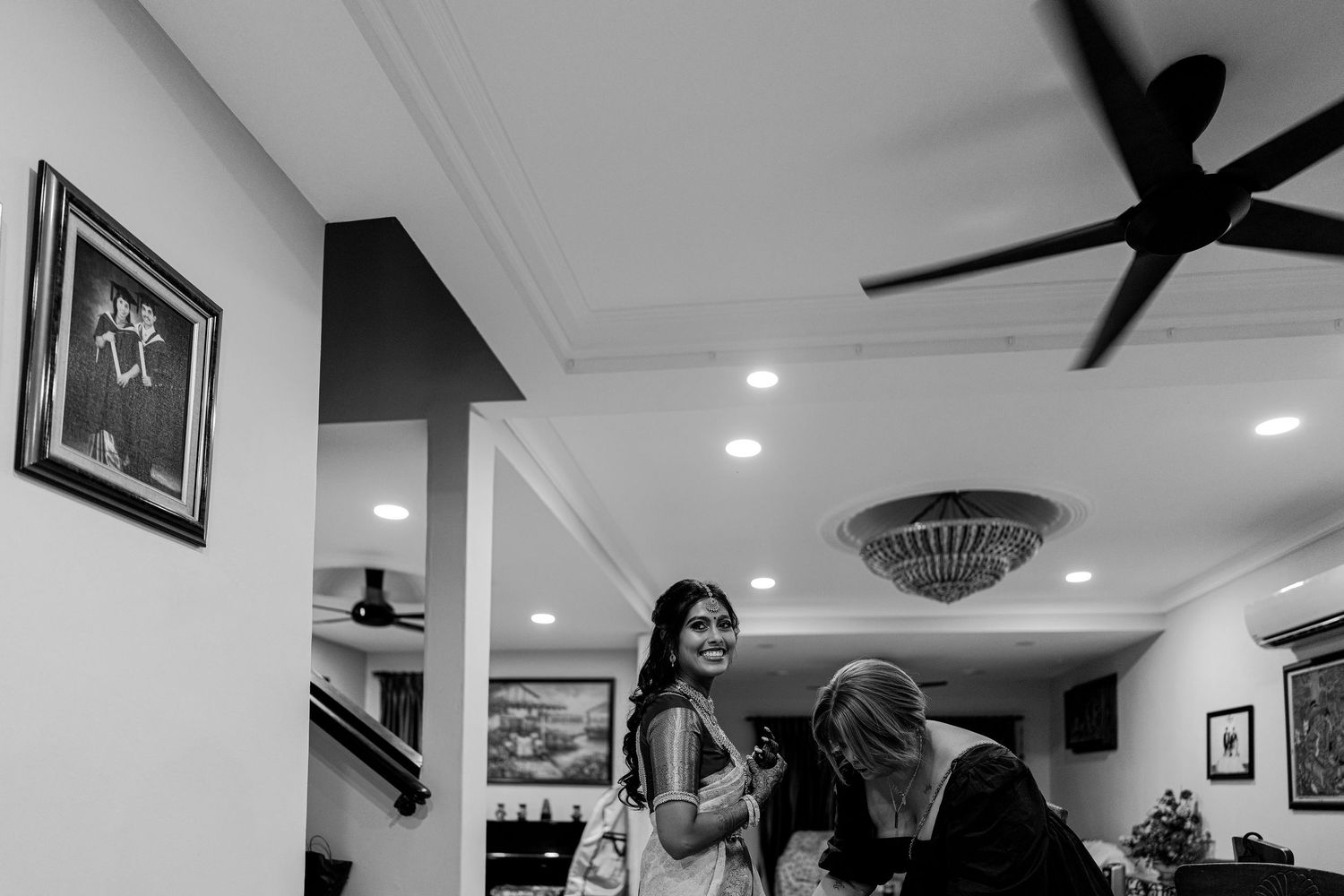 Black and white photo of a smiling woman in traditional attire chatting indoors with ceiling fan above.