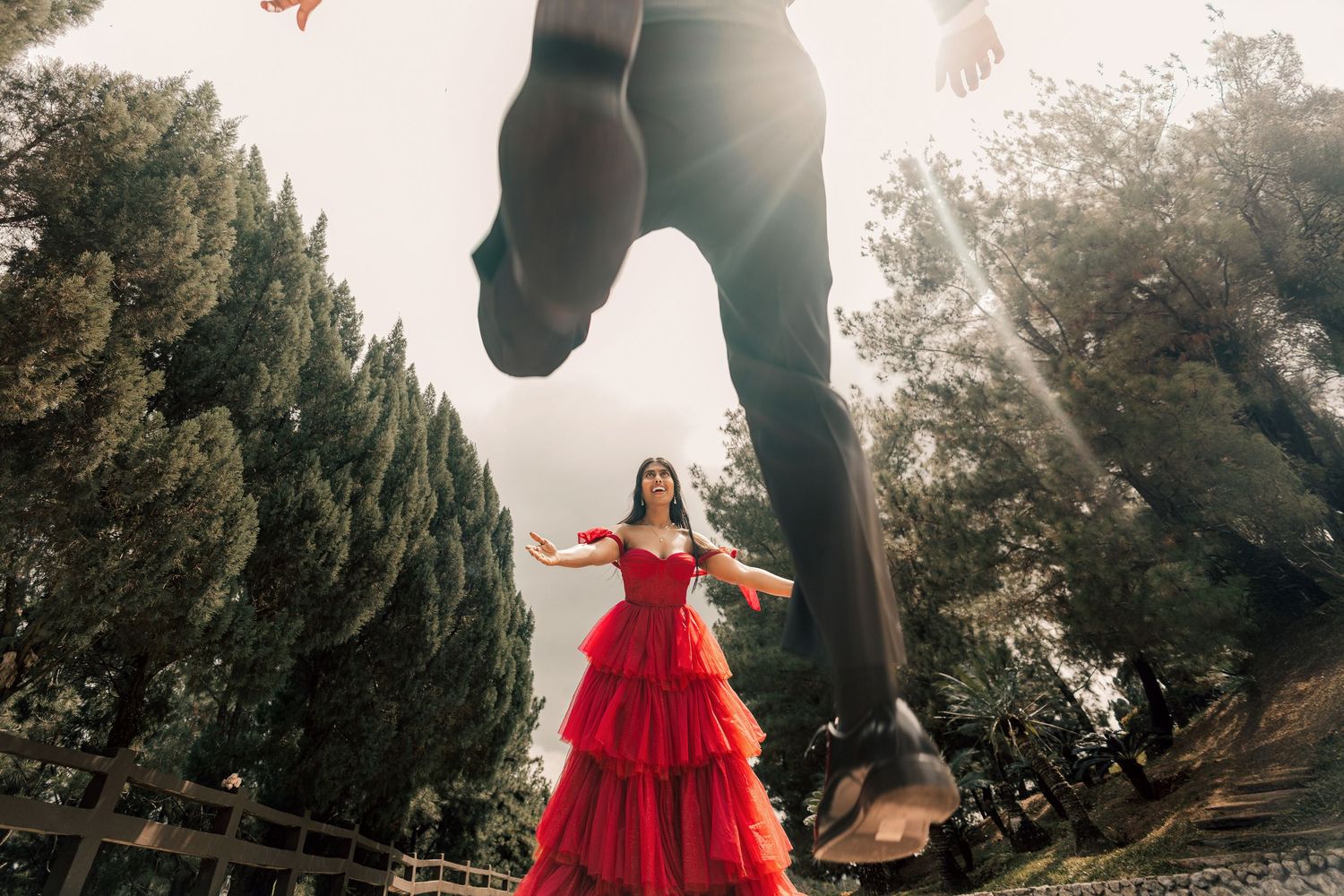 Woman in red ruffled gown poses outdoors while man stands above in creative low-angle wedding photo.