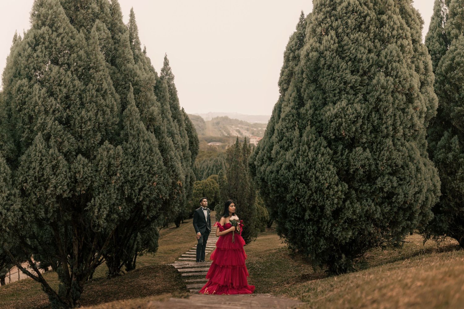 Bride in red gown and groom walk among tall cypress trees on steps at an outdoor wedding venue.