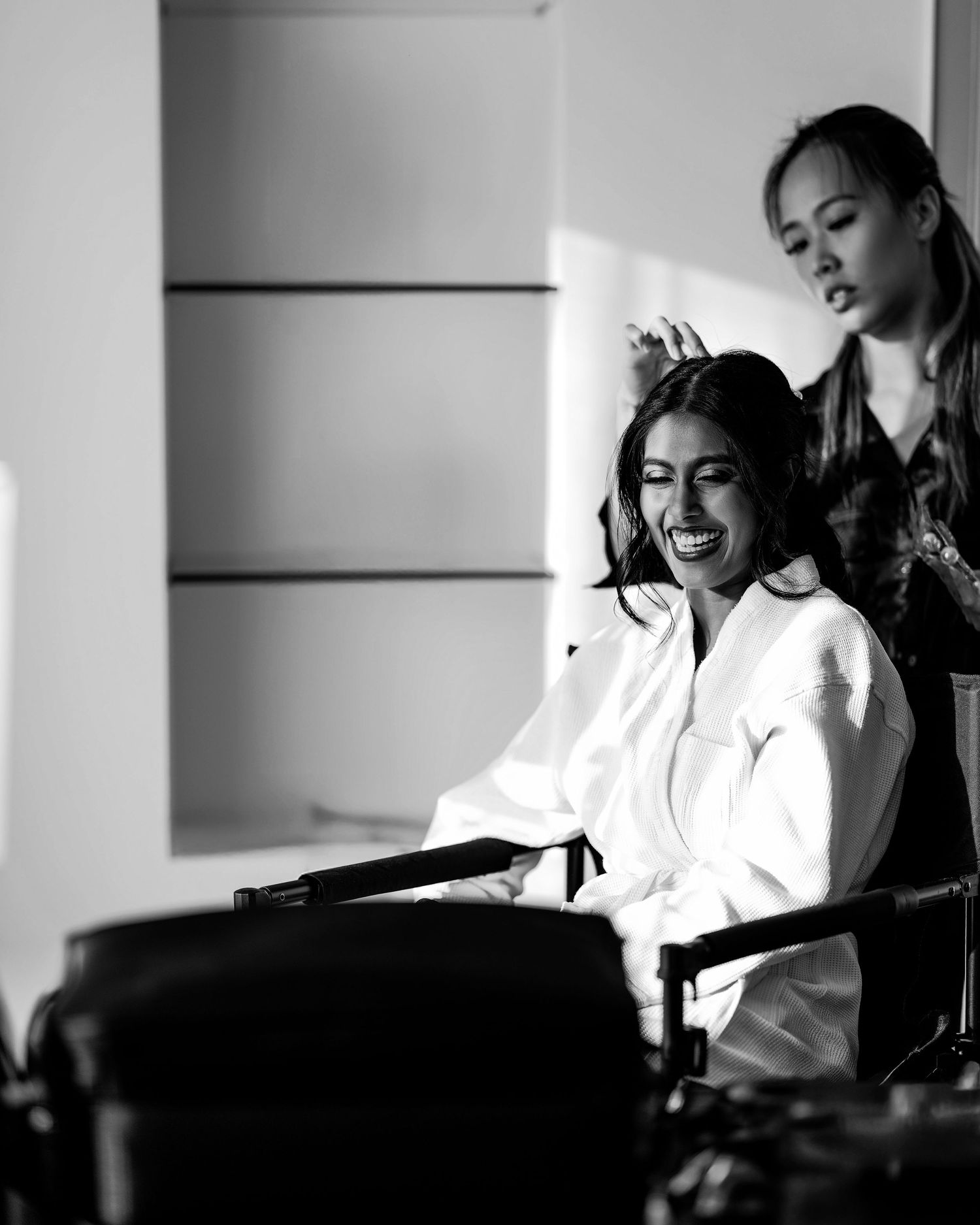 Black and white photo of a smiling woman in a white robe getting her hair done by a stylist.