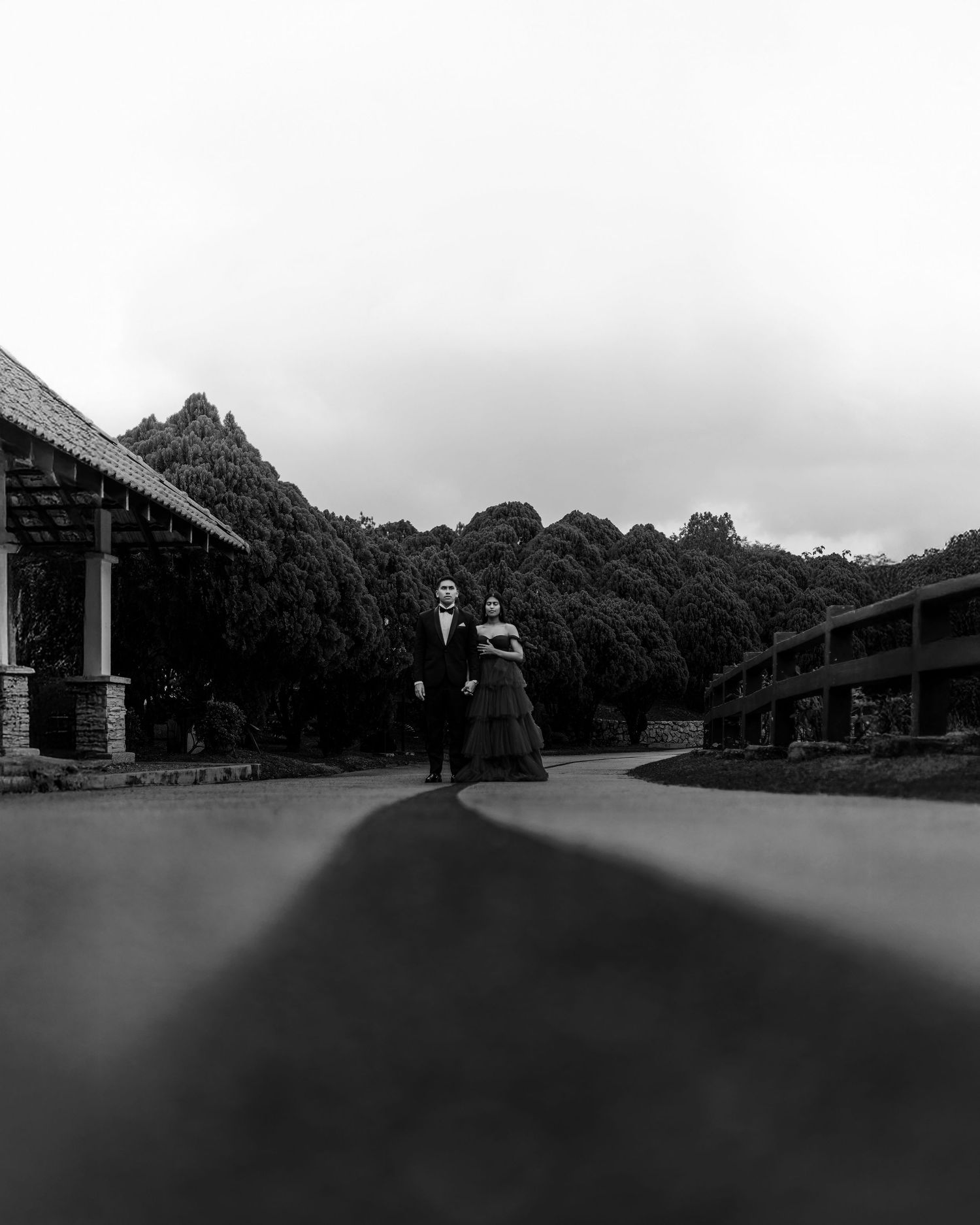 Black and white photo of an elegant couple in formal attire standing on a curved driveway surrounded by lush trees.