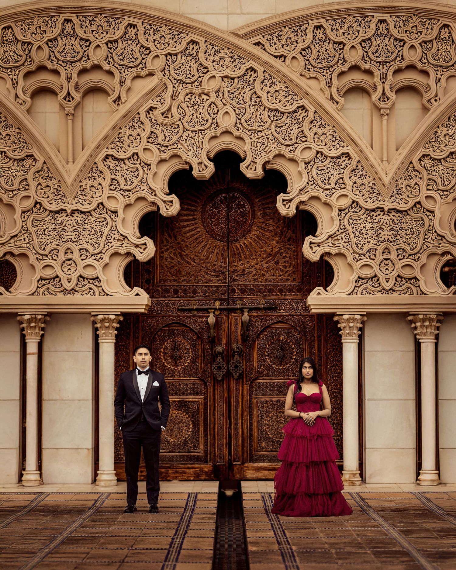 Elegant couple poses in formal attire against ornate Moorish architecture with intricate carved stone details and wooden doors.