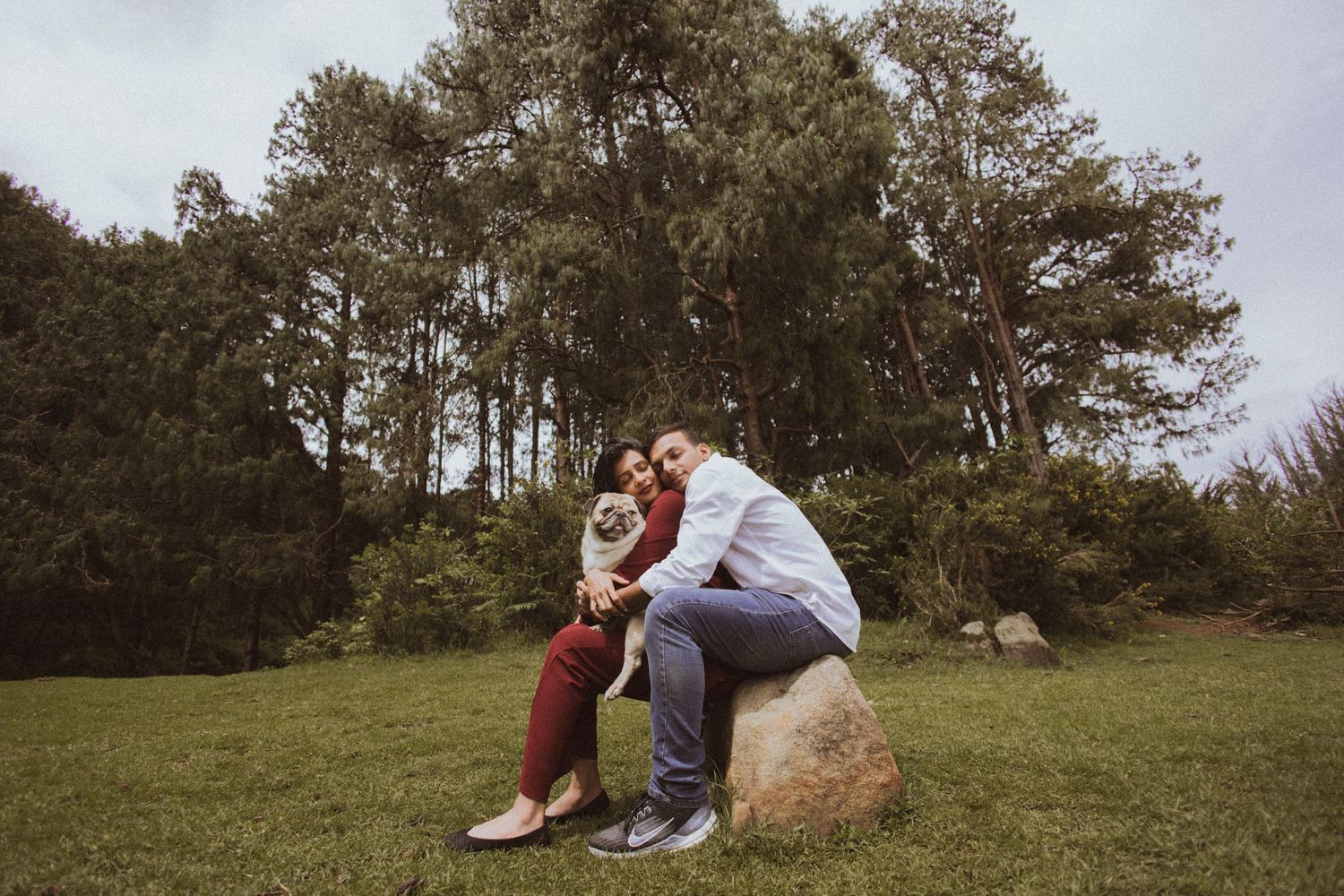 A couple enjoys a peaceful moment together on a grassy hillside surrounded by trees in natural lighting.
