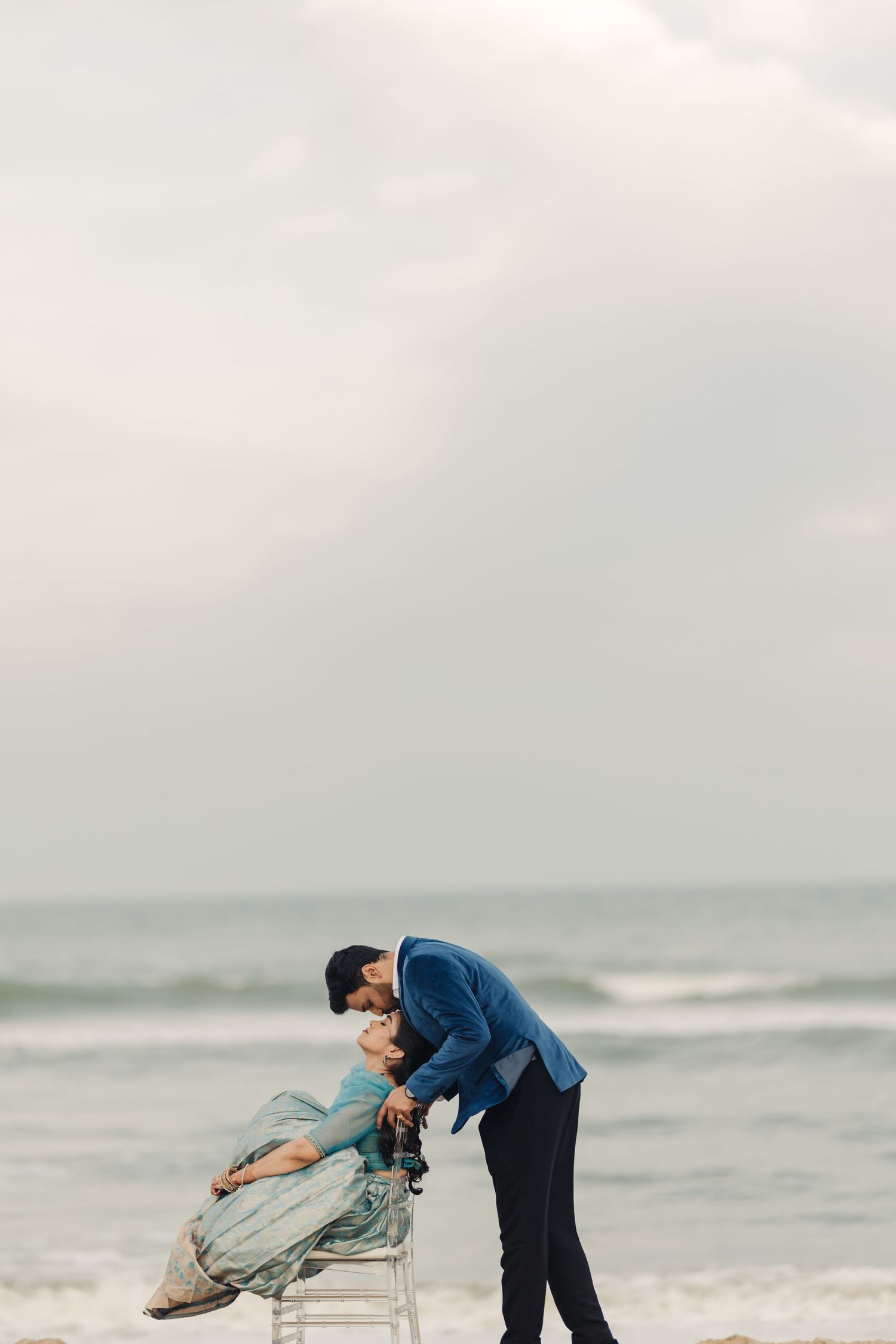 A couple in blue outfits performs a romantic dip pose together on a misty beach shoreline.