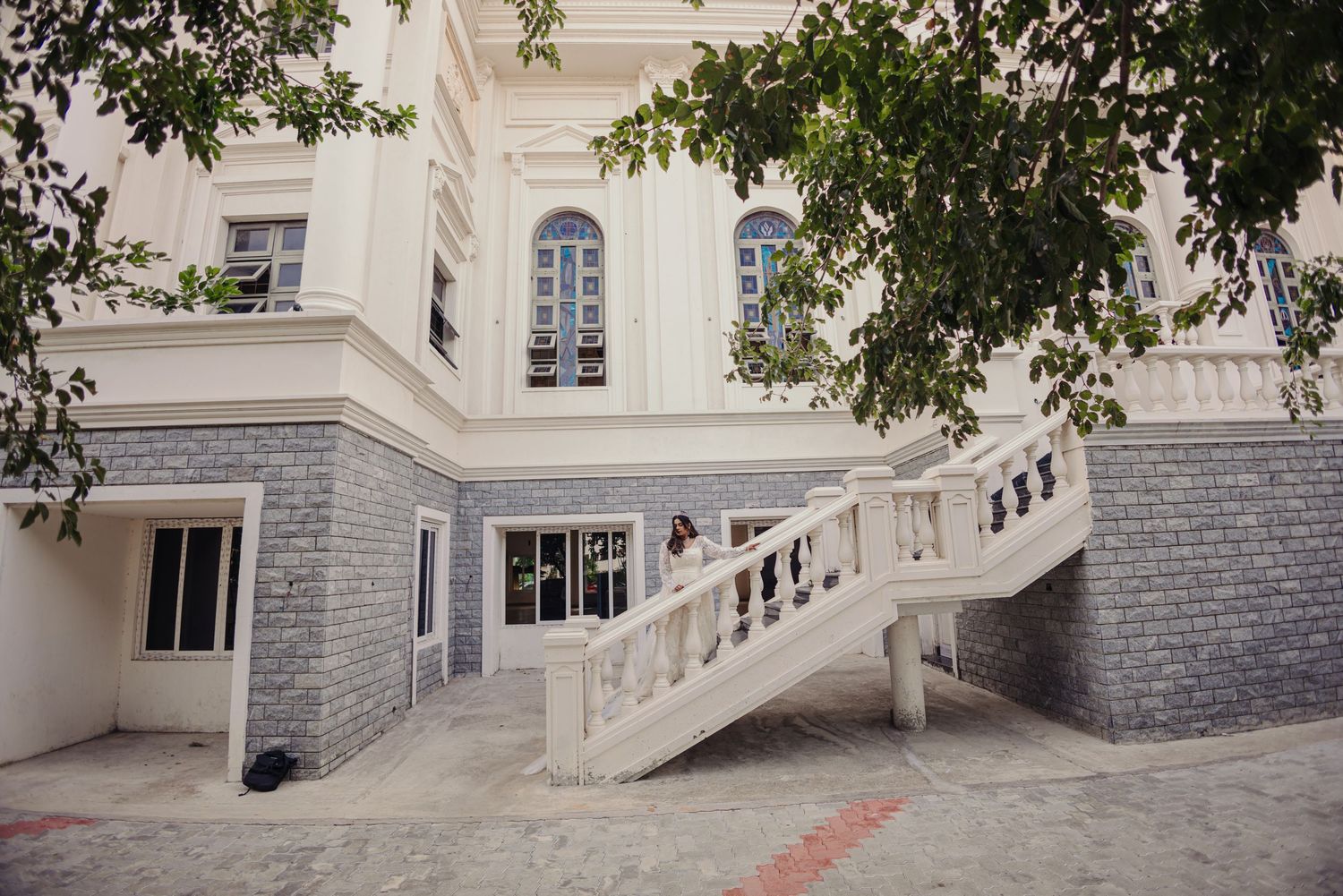 Exterior view of a white building with stone base and ornate staircase surrounded by green trees.