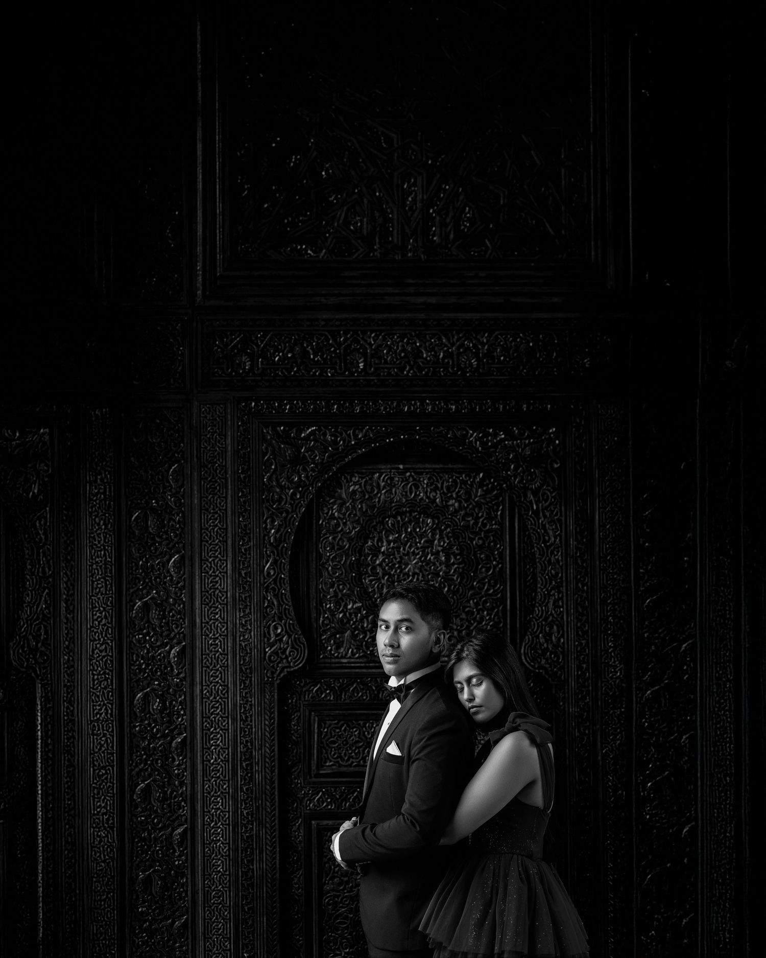 Dramatic black and white portrait of a couple in formal attire posed against an ornate carved dark wall with arched details.
