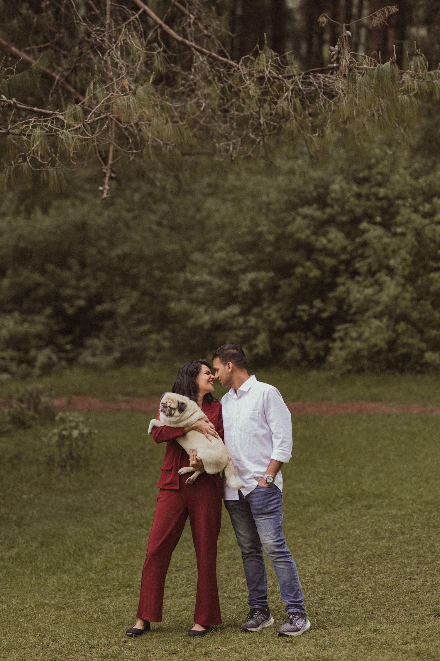 A couple shares a tender moment outdoors while holding their small white dog in a grassy field surrounded by trees.
