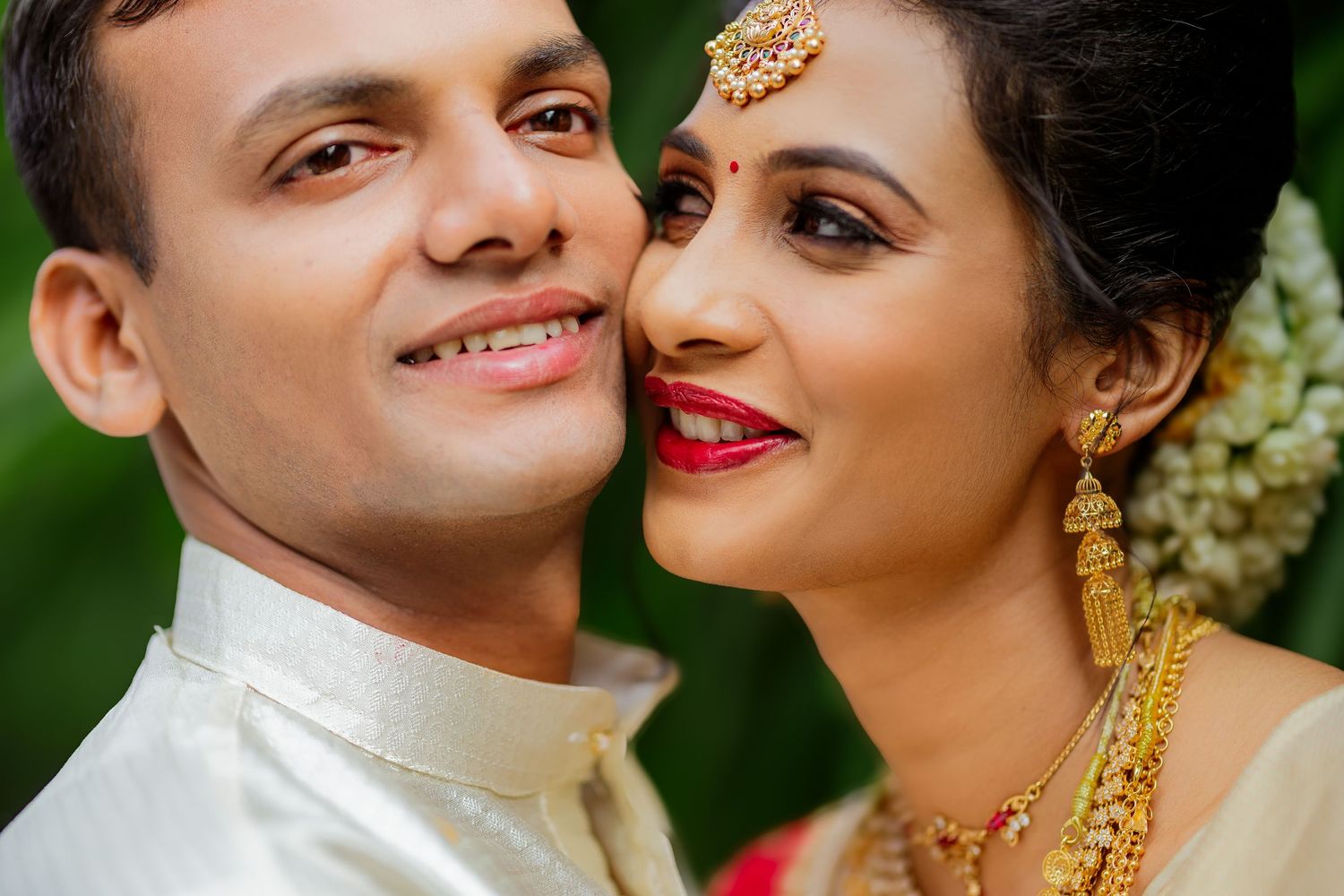 Close up portrait of a couple in traditional Indian wedding attire with gold jewelry against a green background.