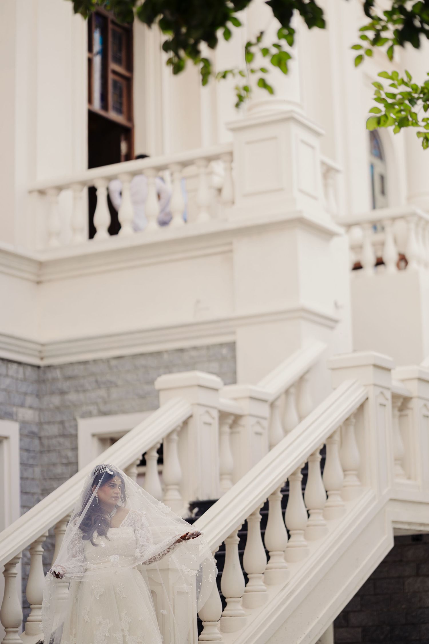 Elegant white wedding dress with lace details flows down ornate stone and marble staircase at historic venue.