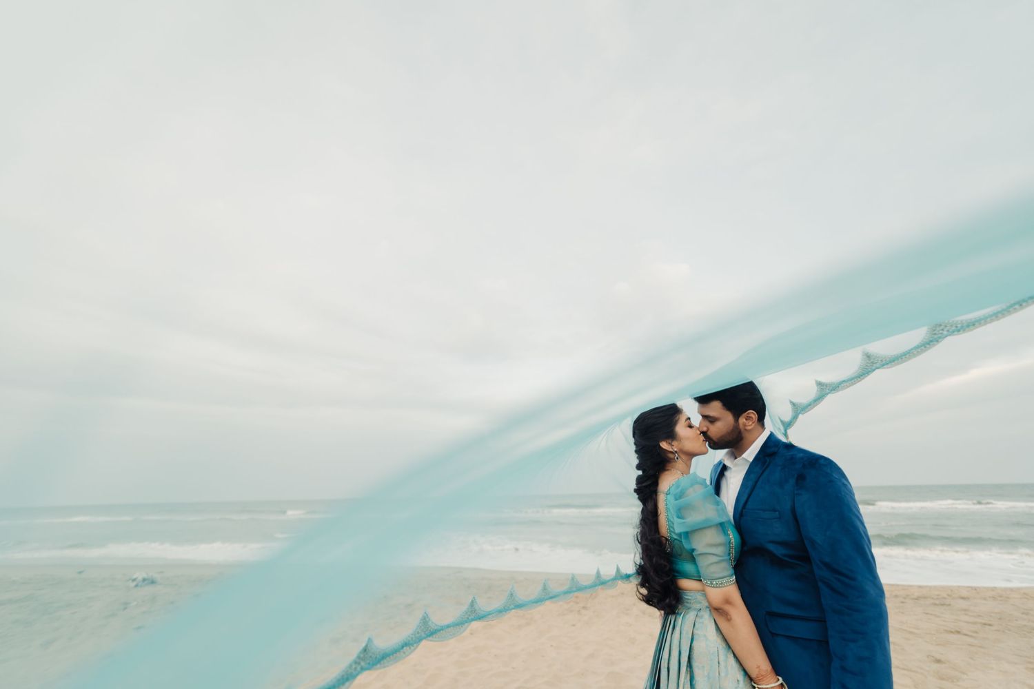 A couple embraces on a misty beach with a flowing blue veil creating an ethereal frame around them.