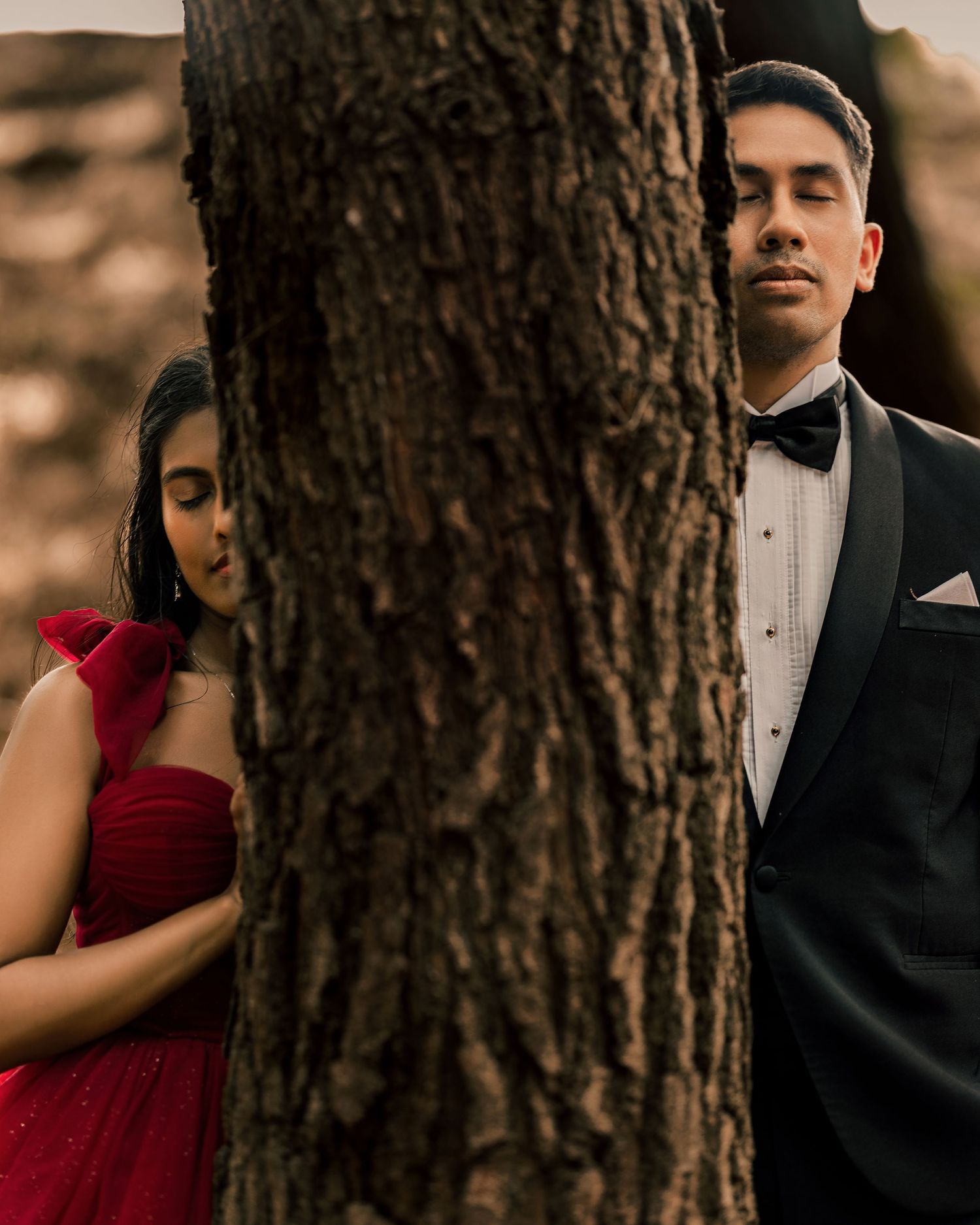 Couple in formal wear posing on opposite sides of a tree, woman in red gown and man in black tuxedo.