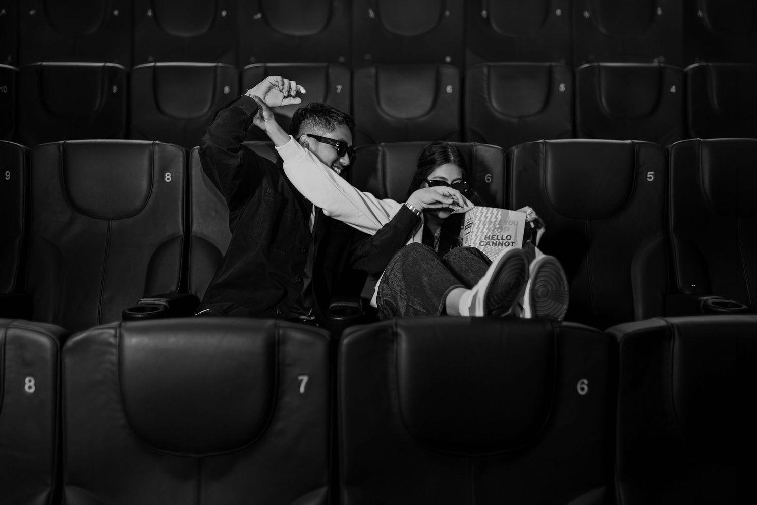 Black and white photo of two people sharing popcorn in empty theater seats.