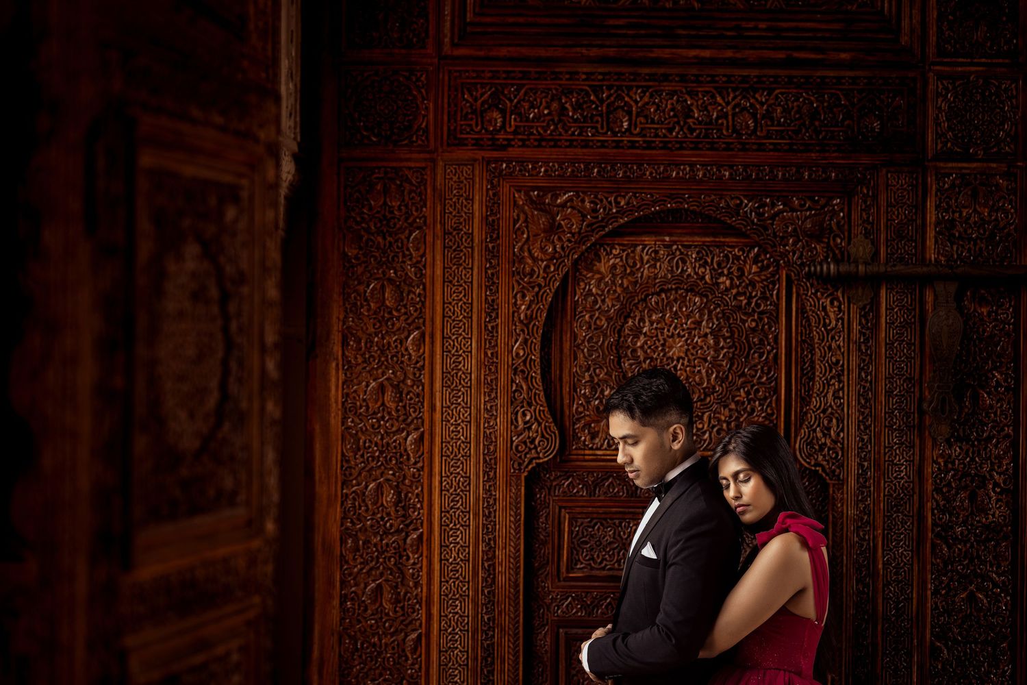 Couple in elegant attire embracing against ornate carved wooden wall with intricate Moroccan patterns.