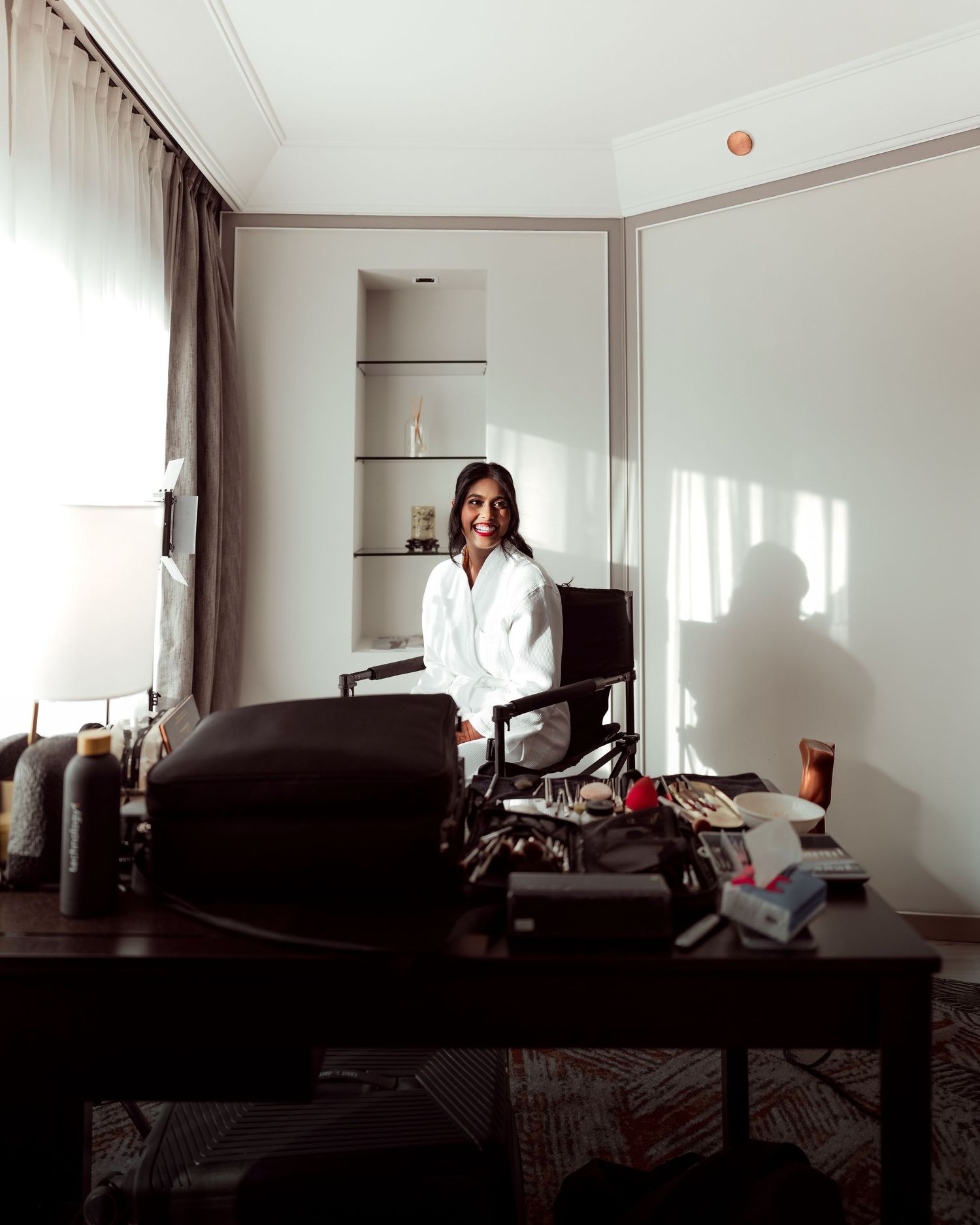Woman in white shirt standing behind a desk in a sunlit hotel room with scattered items on the table.
