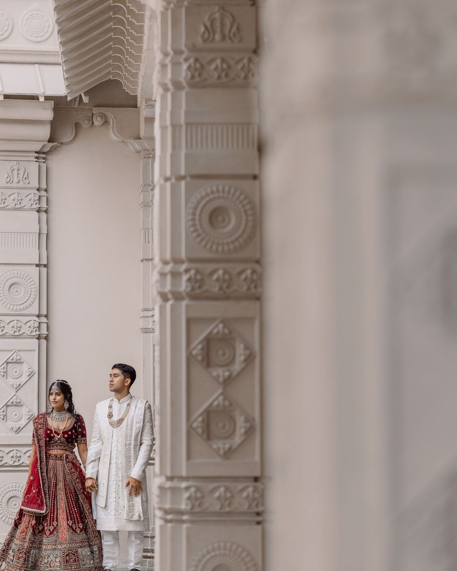 Indian couple in traditional bridal attire holding hands beside ornate white temple columns.