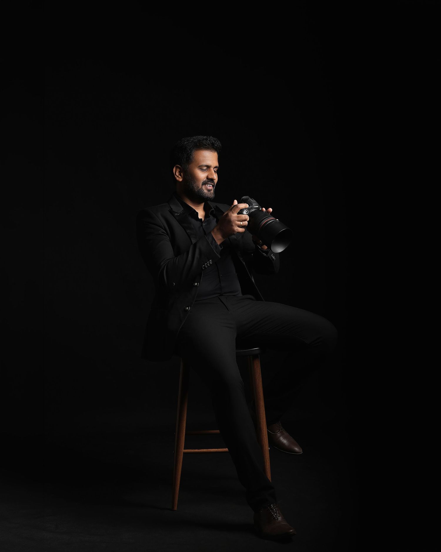 Man in black outfit sitting on stool holding camera against dark background, professional photography portrait.
