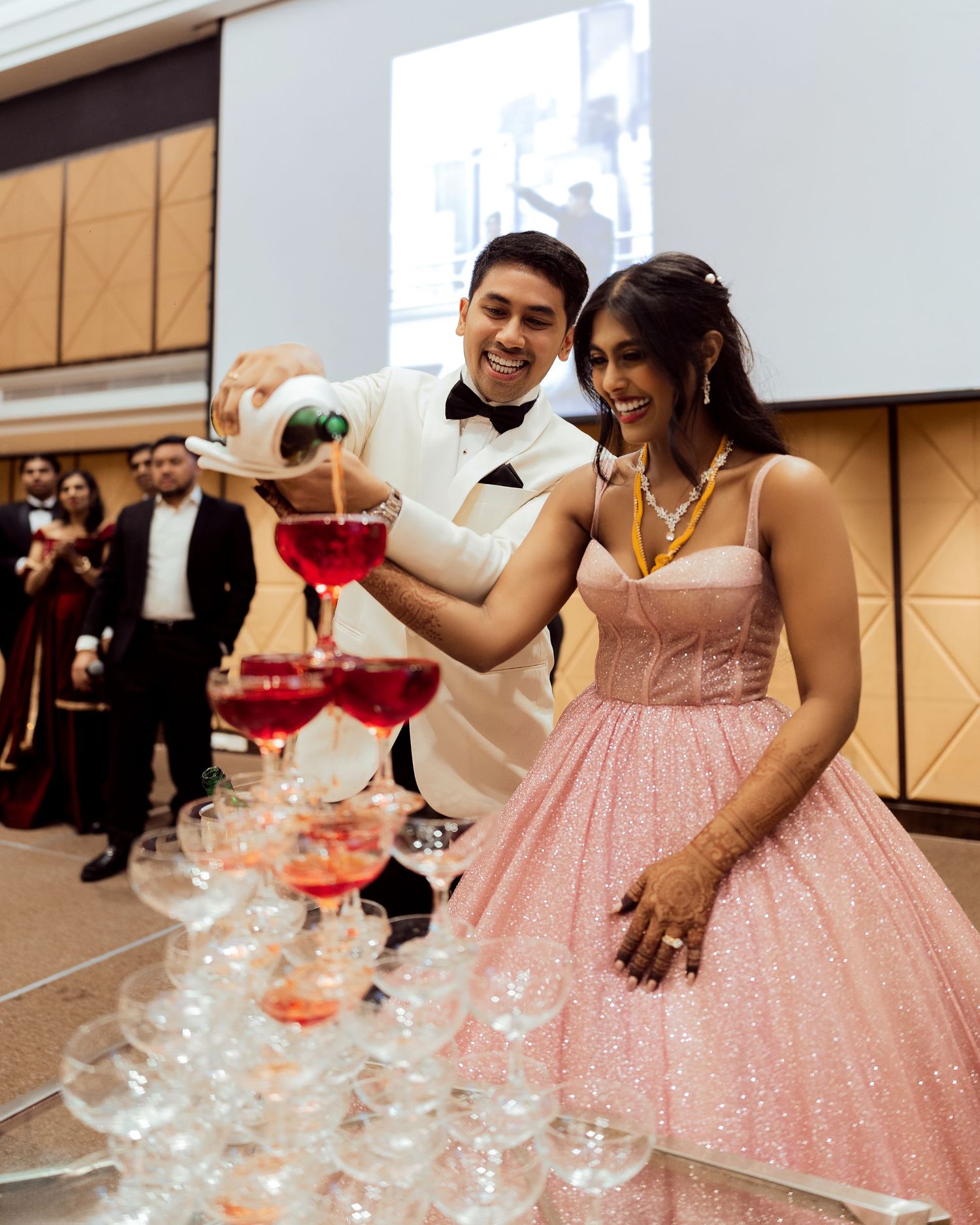 Couple pouring red champagne tower at elegant wedding reception, man in white tuxedo, woman in pink sparkly dress.