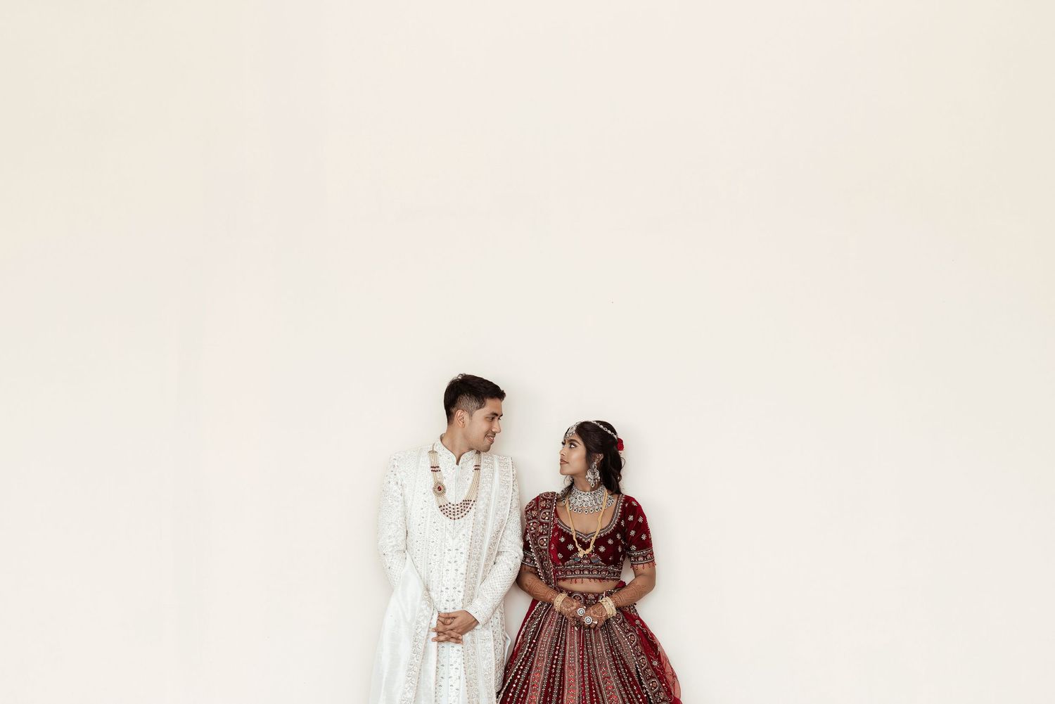 Couple in traditional South Asian wedding attire, man in white sherwani and woman in red lehenga, smiling at each other.
