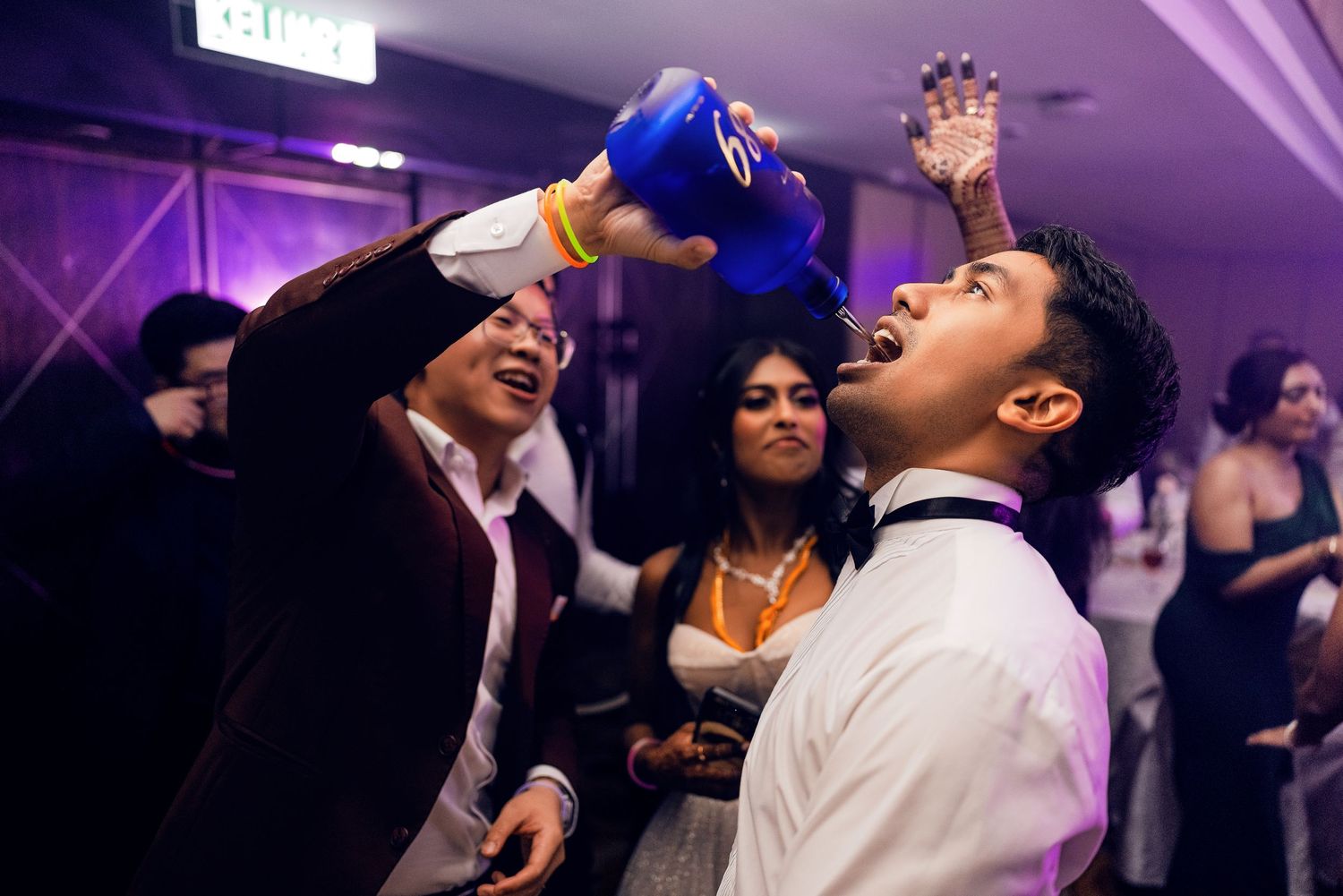 Man pouring drink into another mans mouth at a lively party event with guests dancing in background.