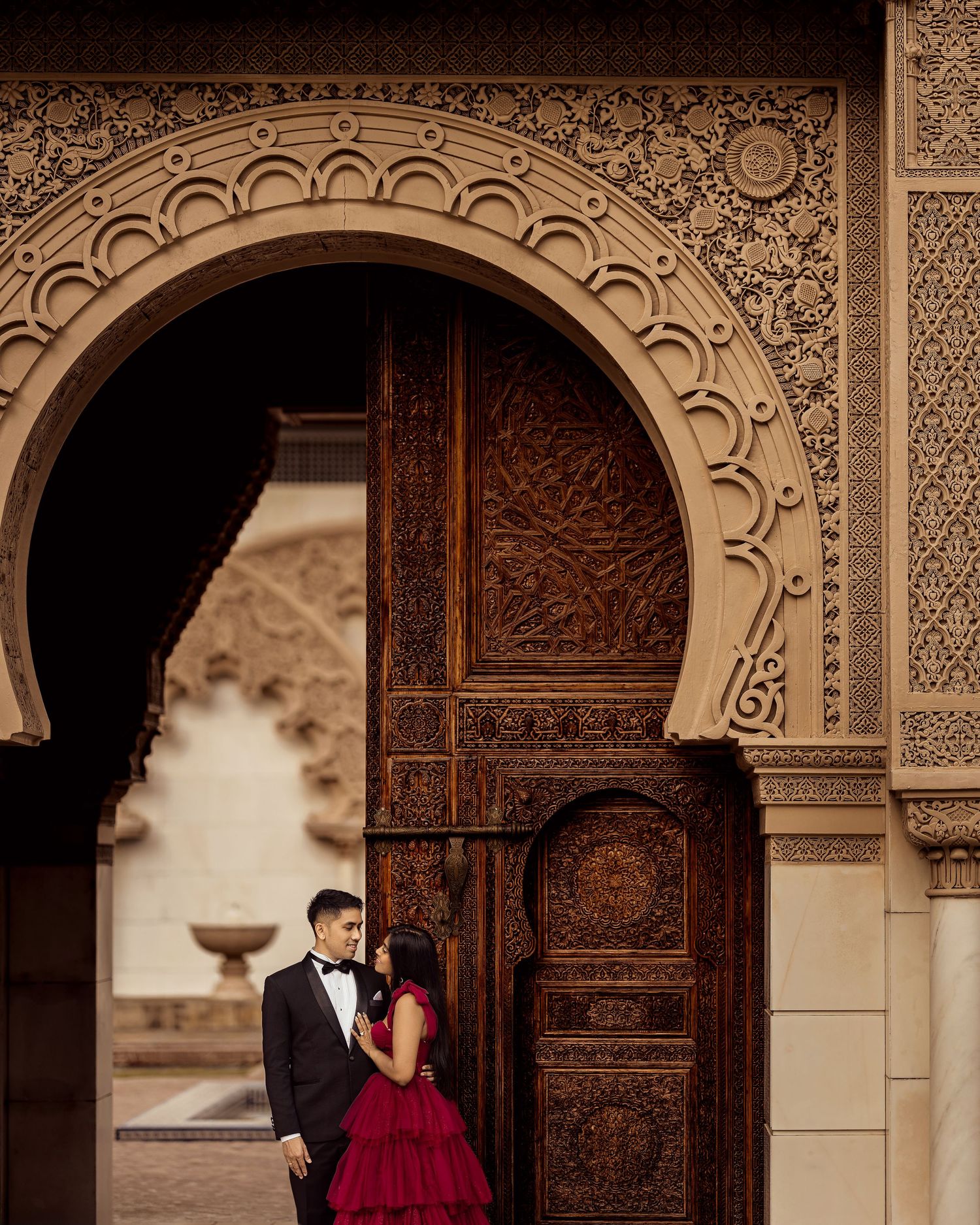 Couple in formal attire embracing under ornate Moroccan archway with intricate carved wooden doors.