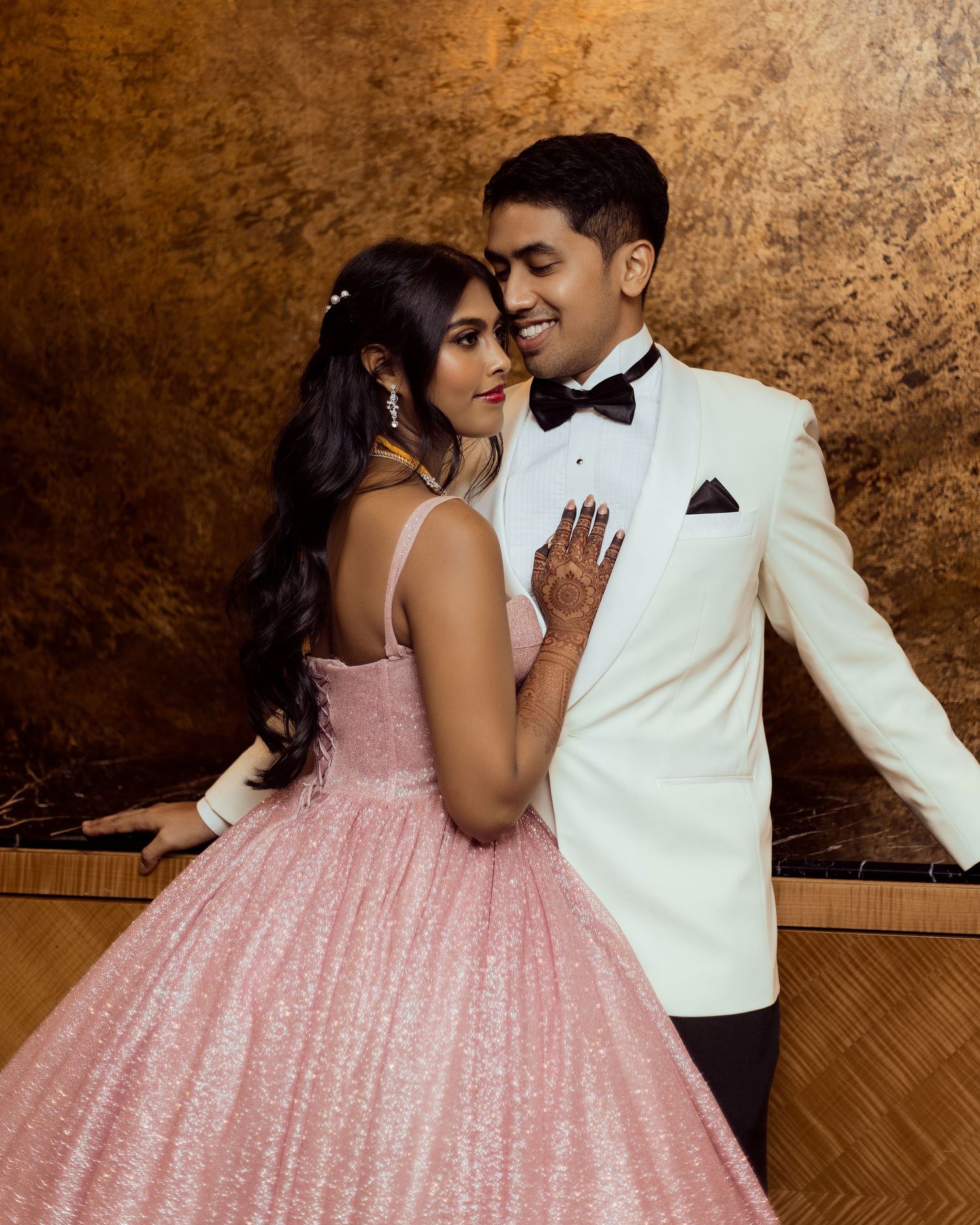 A couple poses elegantly, woman in pink glitter ball gown and man in white tuxedo, sharing a romantic moment.