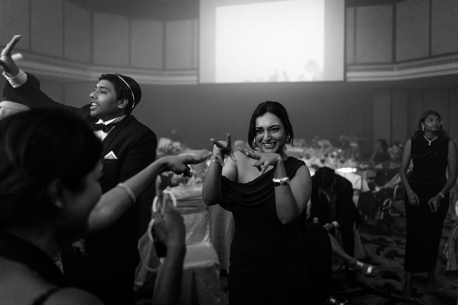 Black and white photo of a woman smiling and clapping at an elegant indoor event with guests in formal attire.