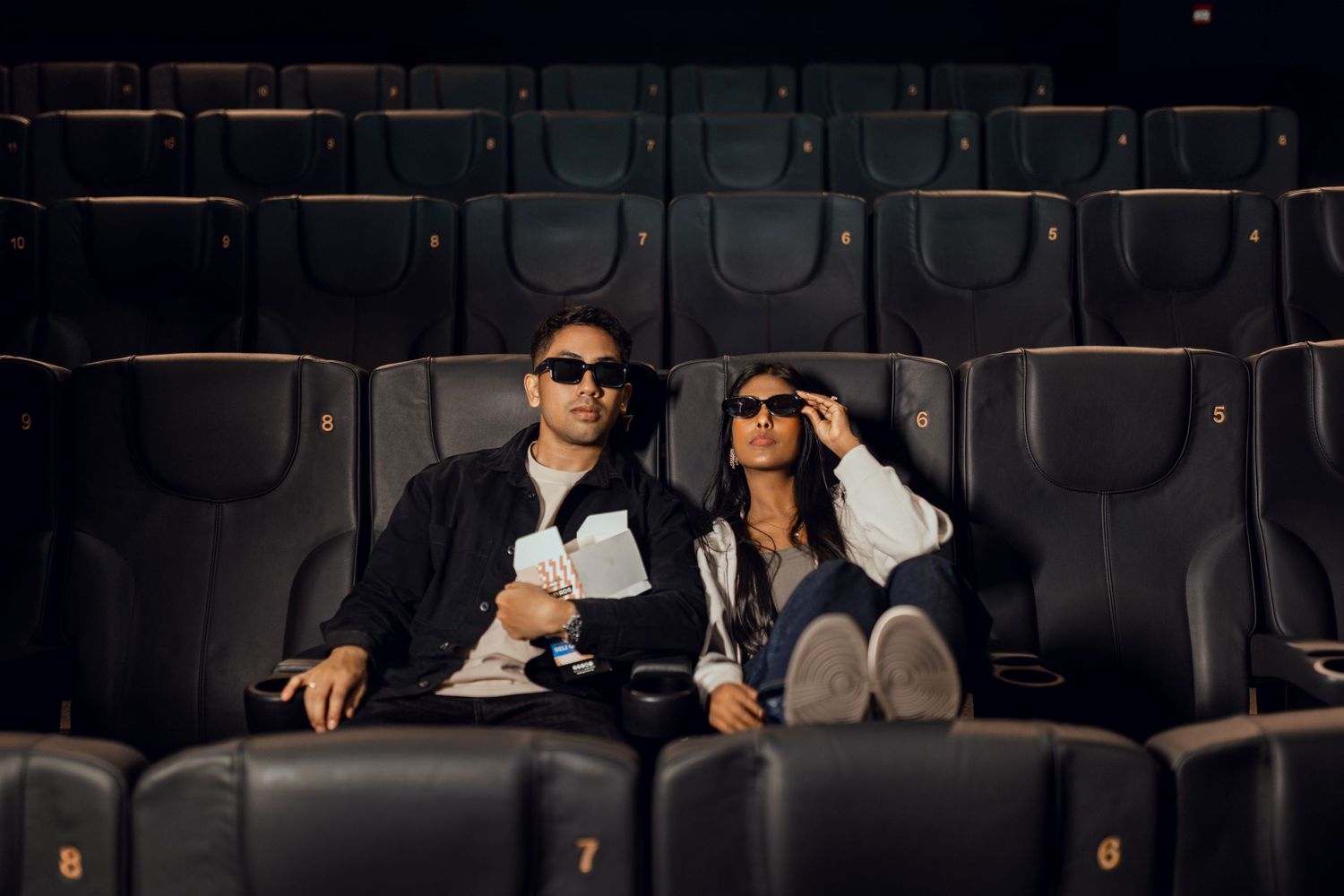 Couple wearing 3D glasses watching a movie together in a dark cinema with rows of empty seats behind them.