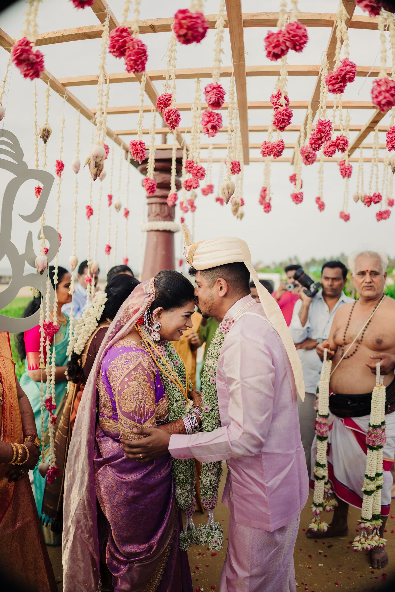 Traditional Indian wedding ceremony under a canopy decorated with hanging pink floral arrangements and garlands.