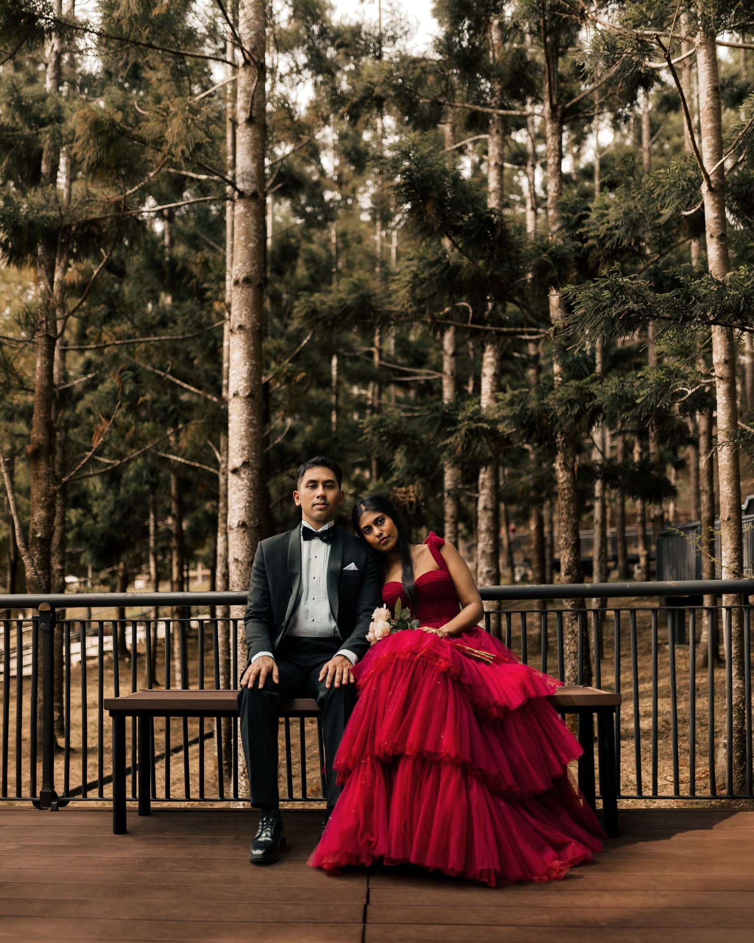 Couple in formal attire posing on bench in pine forest, man in tuxedo and woman in red ball gown.
