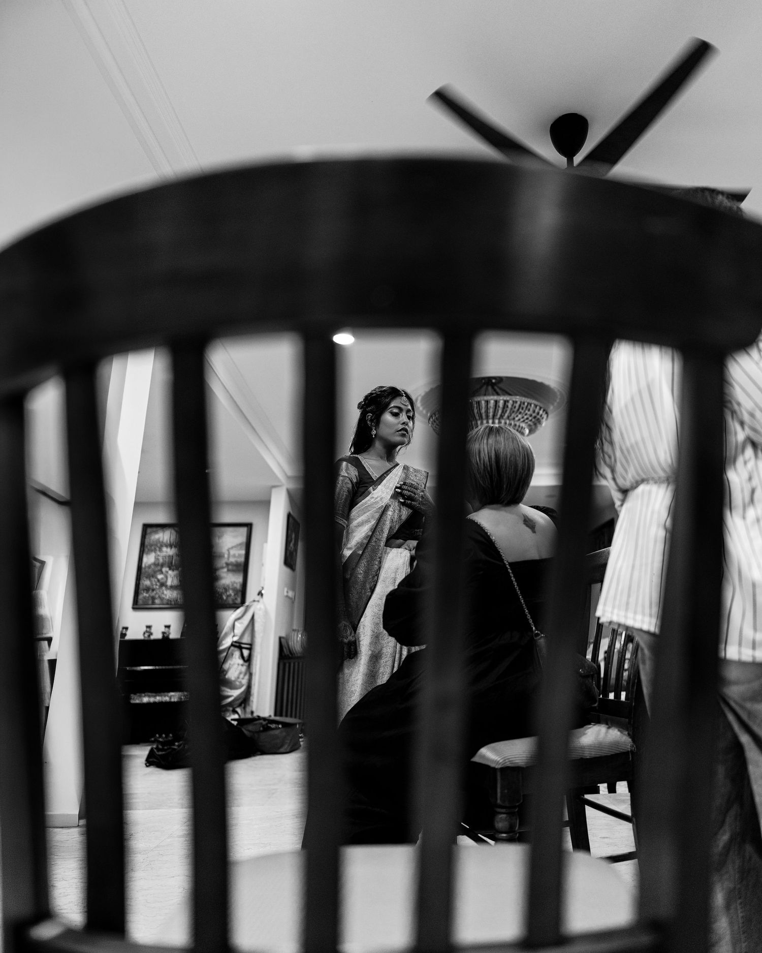 Black and white photo of a woman in a saree viewed through chair slats during a wedding preparation.